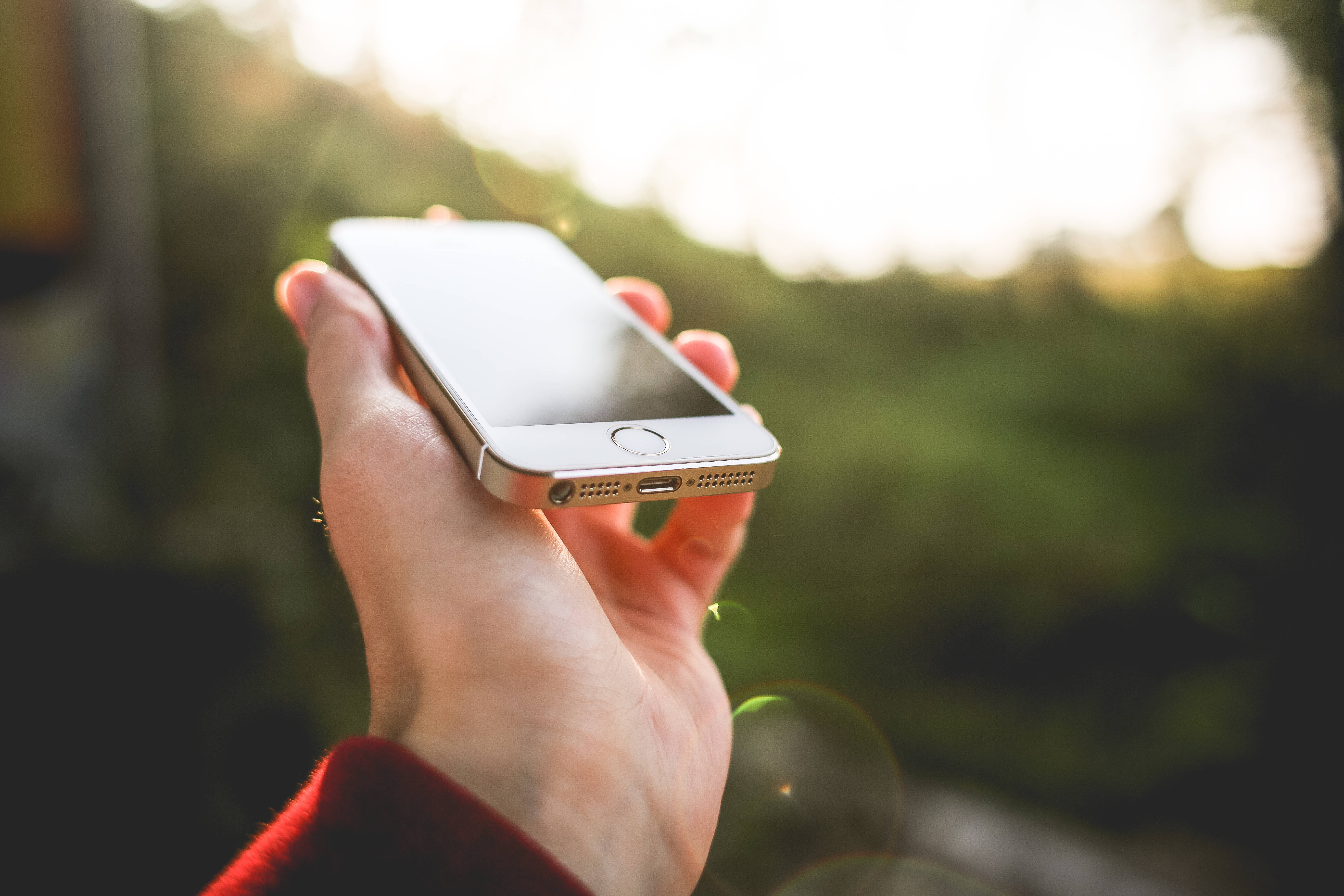 original iPhone 5 桌布 壁紙 — 一隻手拿著銀色智慧型手機，背景是陽光照射下模糊的綠色和鏡頭光暈。