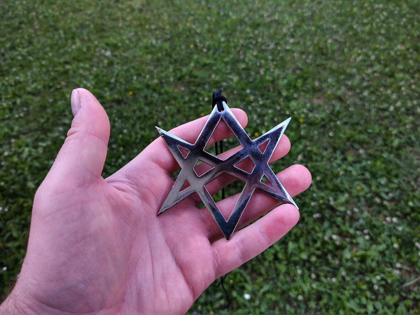 Thelema 桌布 壁紙 — 一隻手拿著閃亮的銀色五角星吊墜，背景是模糊的綠色草地。