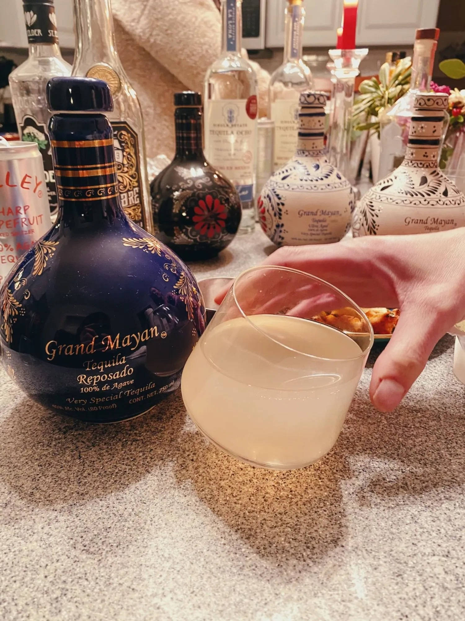 alcohol 桌布 壁紙 — 一隻手拿著雞尾酒杯，旁邊放著一瓶藍色Grand Mayan龍舌蘭酒，在斑點檯面上還有其他烈酒和小吃。