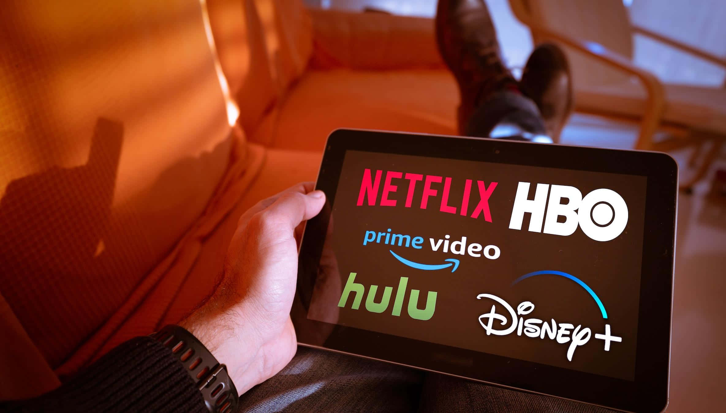 HBO 桌布 壁紙 — 一隻手拿著顯示串流服務標誌的平板電腦，在橙色沙發上放鬆，雙腳翹起。