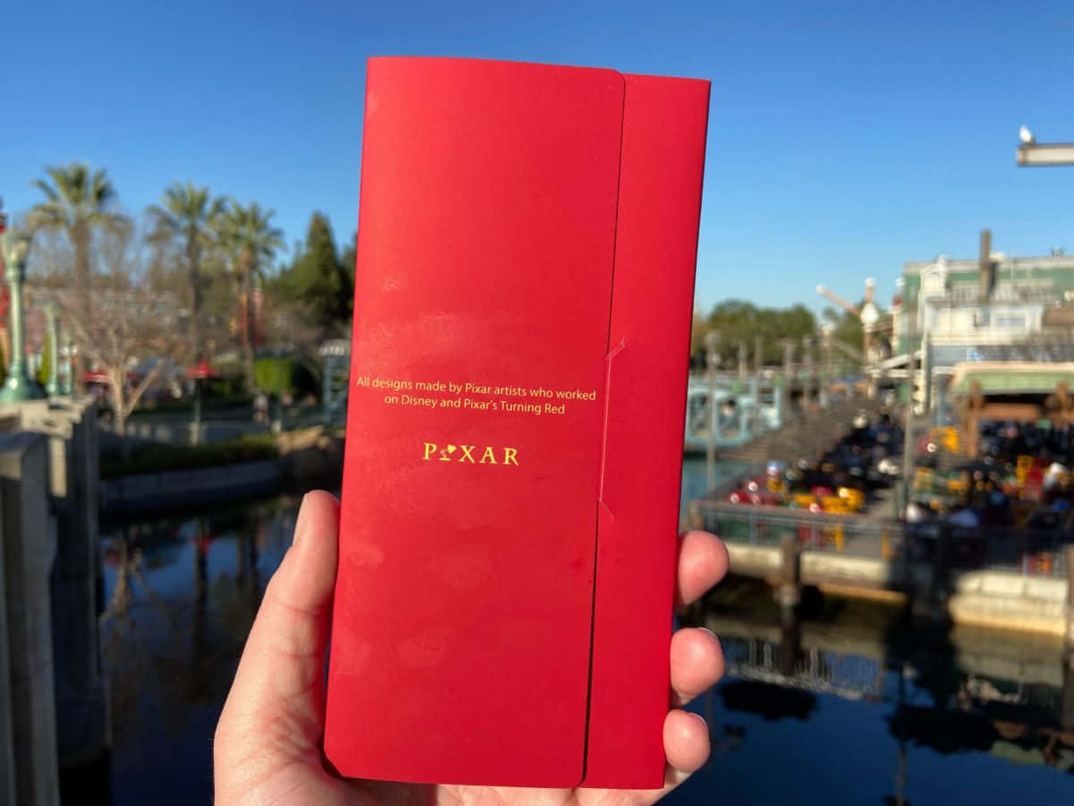 Red Envelope 桌布 壁紙 — 一隻手拿著鮮紅色的Pixar小冊子，背景是陽光明媚的迪士尼樂園，有棕櫚樹和水。