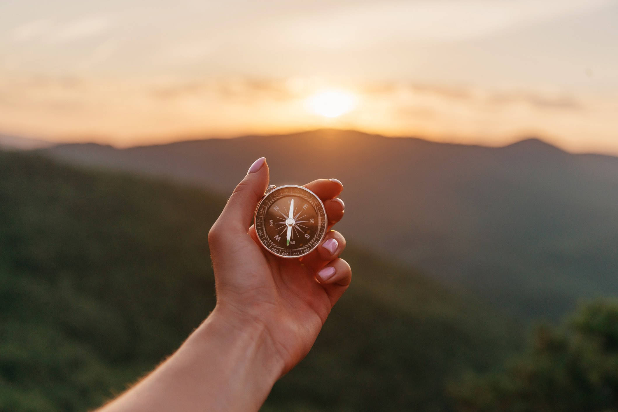 日落 桌布 壁紙 — 一隻手拿著黃銅指南針，背景是金色夕陽下的連綿綠色山丘。