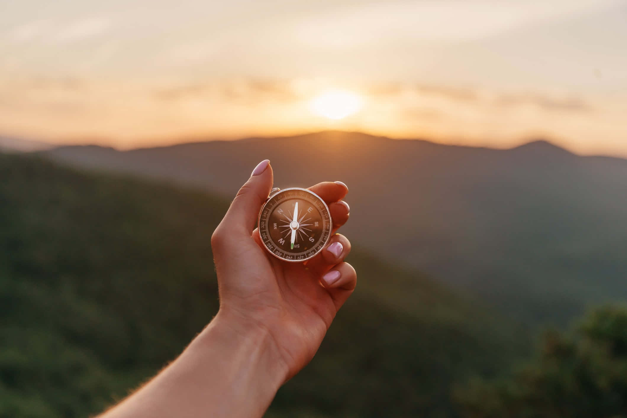 life 桌布 壁紙 — 一隻手拿著黃銅指南針，背景是金色夕陽下的連綿綠色山丘。