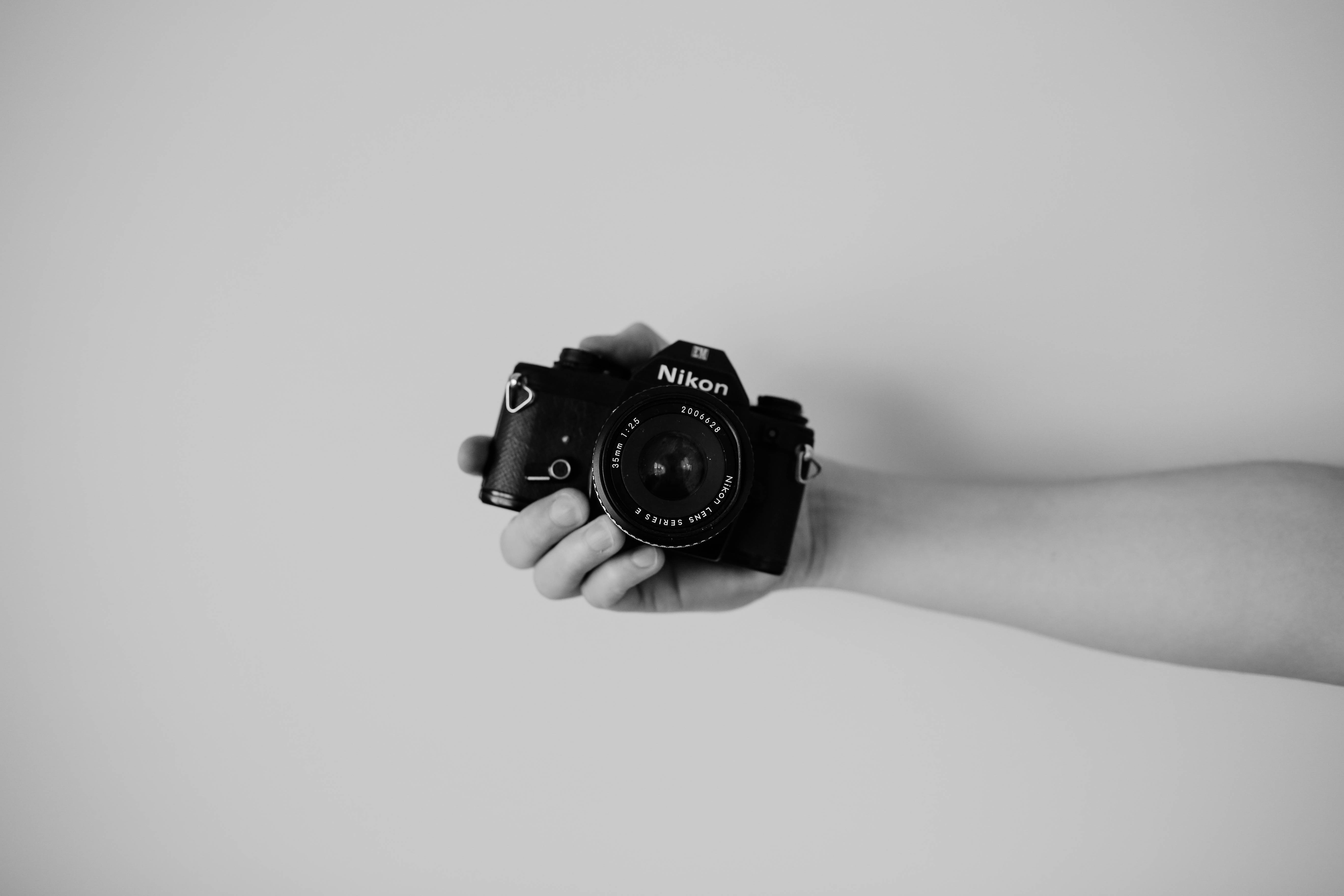 攝影 桌布 壁紙 — 一隻手拿著黑色 Nikon 相機，背景是單色的淺灰色。