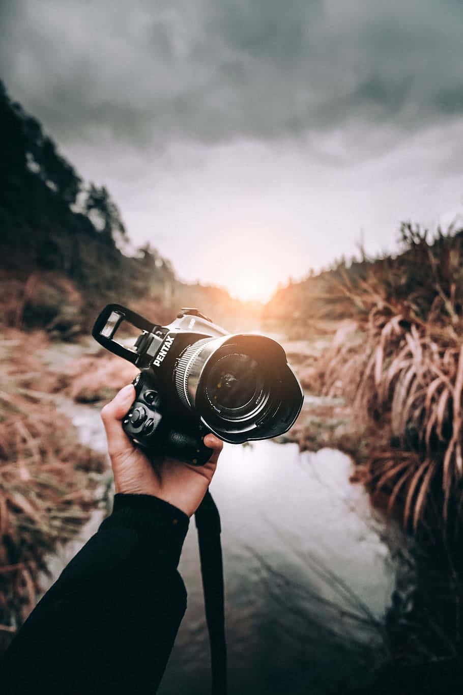 Dslr Camera 桌布 壁紙 — 一隻手拿著黑色 Pentax 相機，在陰鬱的雲層下框住陽光明媚的河谷。