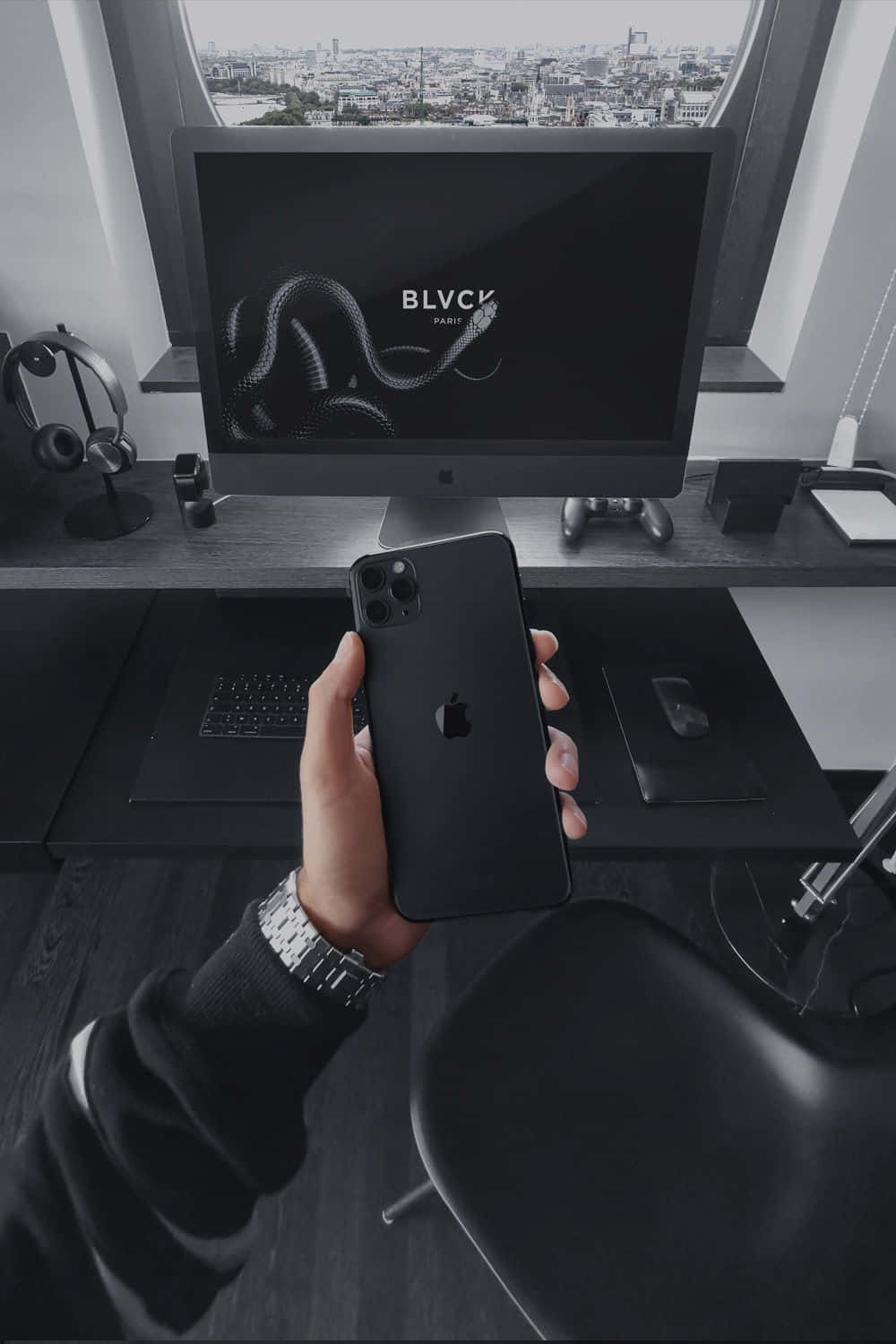 黑色 桌布 壁紙 — 一隻手拿著黑色iPhone，面前是時尚的辦公桌設置，顯示器上顯示蛇形標誌和窗外城市景觀。