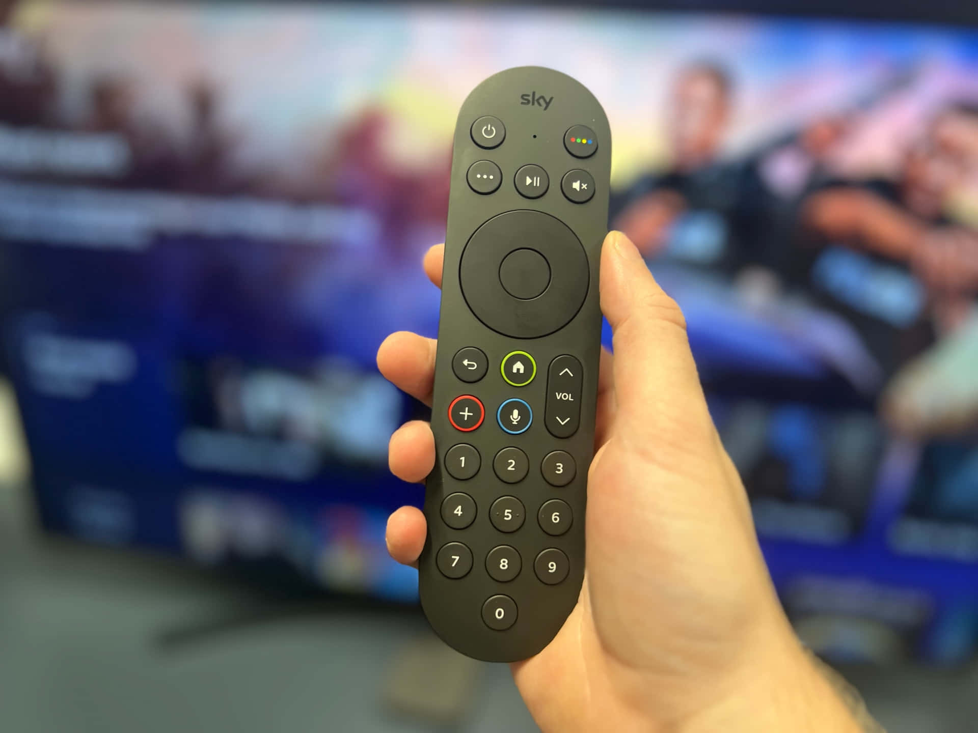watching tv 桌布 壁紙 — 一隻手拿著黑色Sky遙控器，焦點清晰，背景是模糊的電視螢幕，顯示體育賽事轉播。