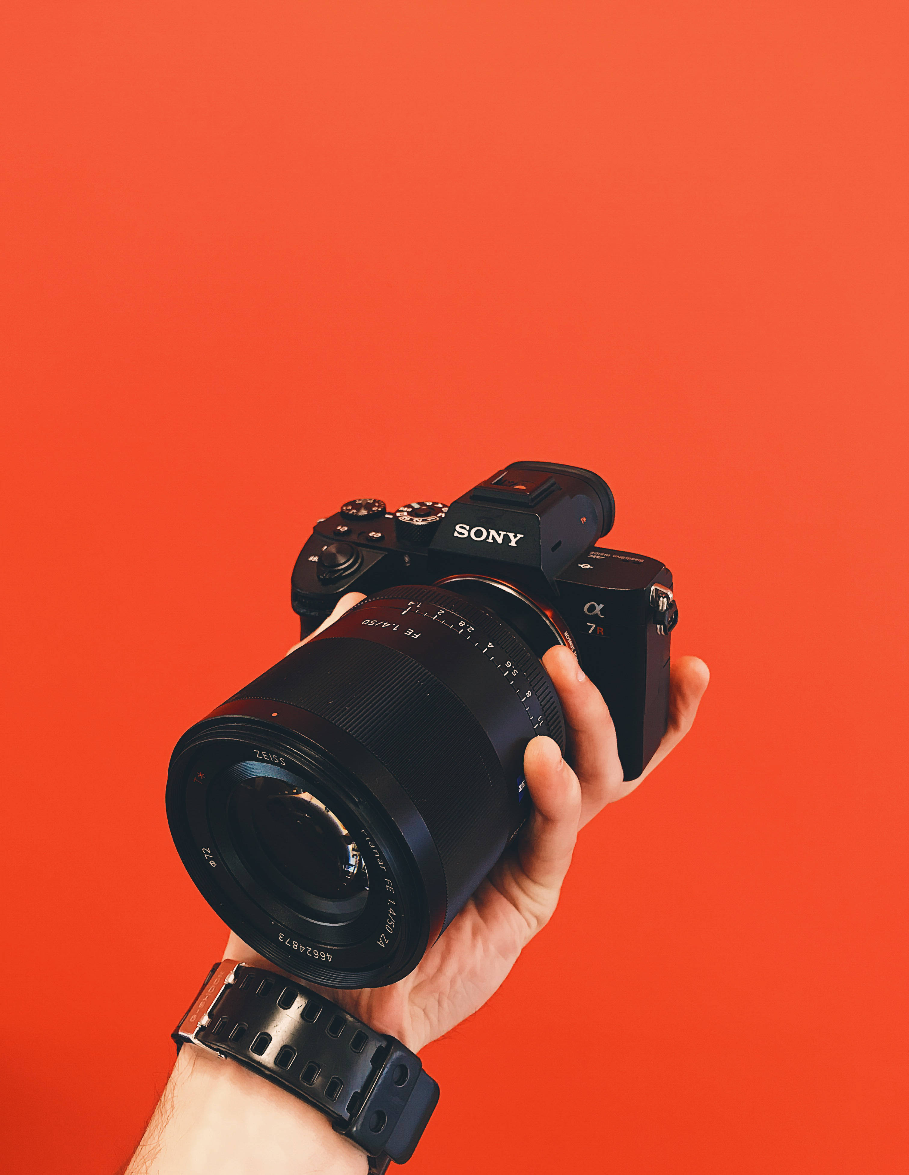 Sony 桌布 壁紙 — 一隻手拿著黑色Sony Alpha 7R相機，背景為鮮豔的橙色。