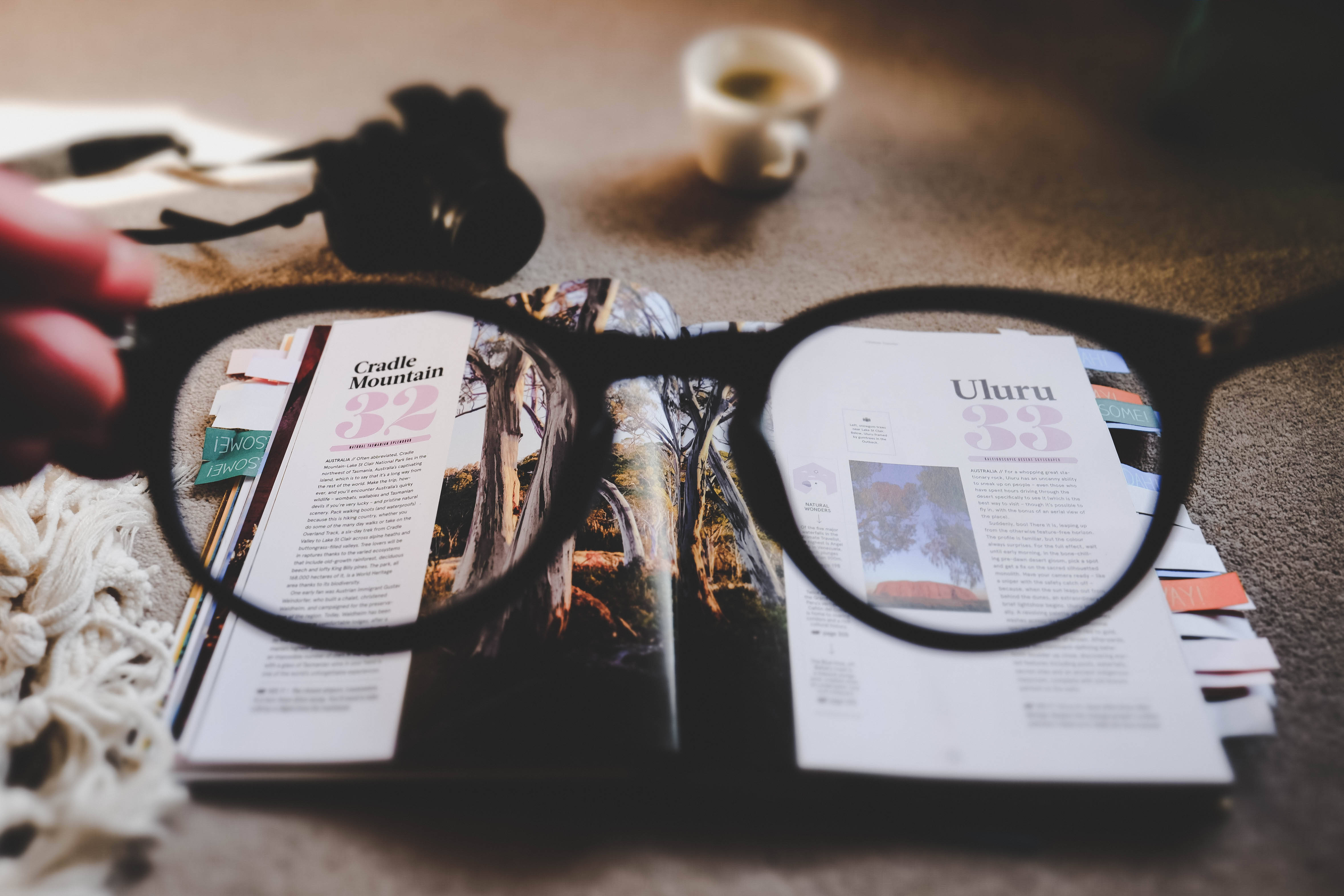 Magazine 桌布 壁紙 — 一隻手拿著黑色框眼鏡，放在打開的旅遊雜誌上，雜誌上有搖籃山和烏魯魯，附近有一杯咖啡。