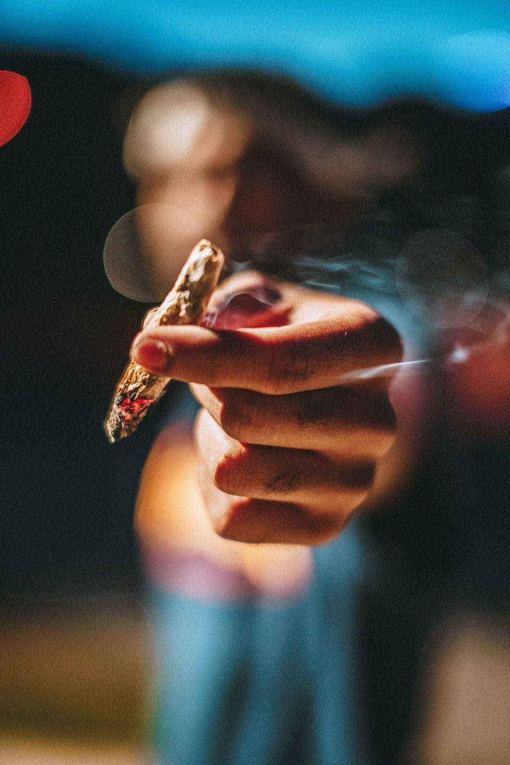 Blunt 桌布 壁紙 — 一隻手拿著點燃的香煙，在模糊的背景和散景燈光中散發煙霧。