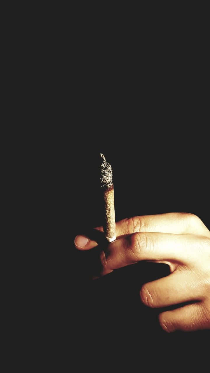 joint 桌布 壁紙 — 一隻手拿著點燃的香煙，背景為純黑色，戲劇性的燈光凸顯了煙霧和手指。