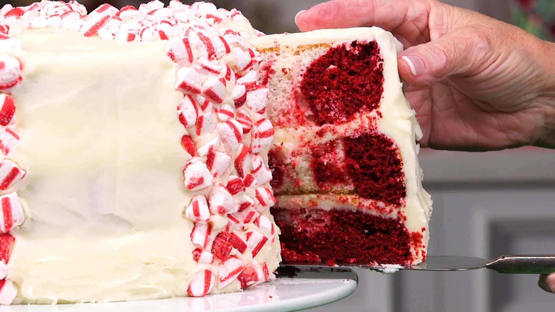 Red Velvet Cake 桌布 壁紙 — 一隻手拿起一片紅絲絨蛋糕，上面有白色糖霜和拐杖糖裝飾，露出濕潤的深紅色層次。