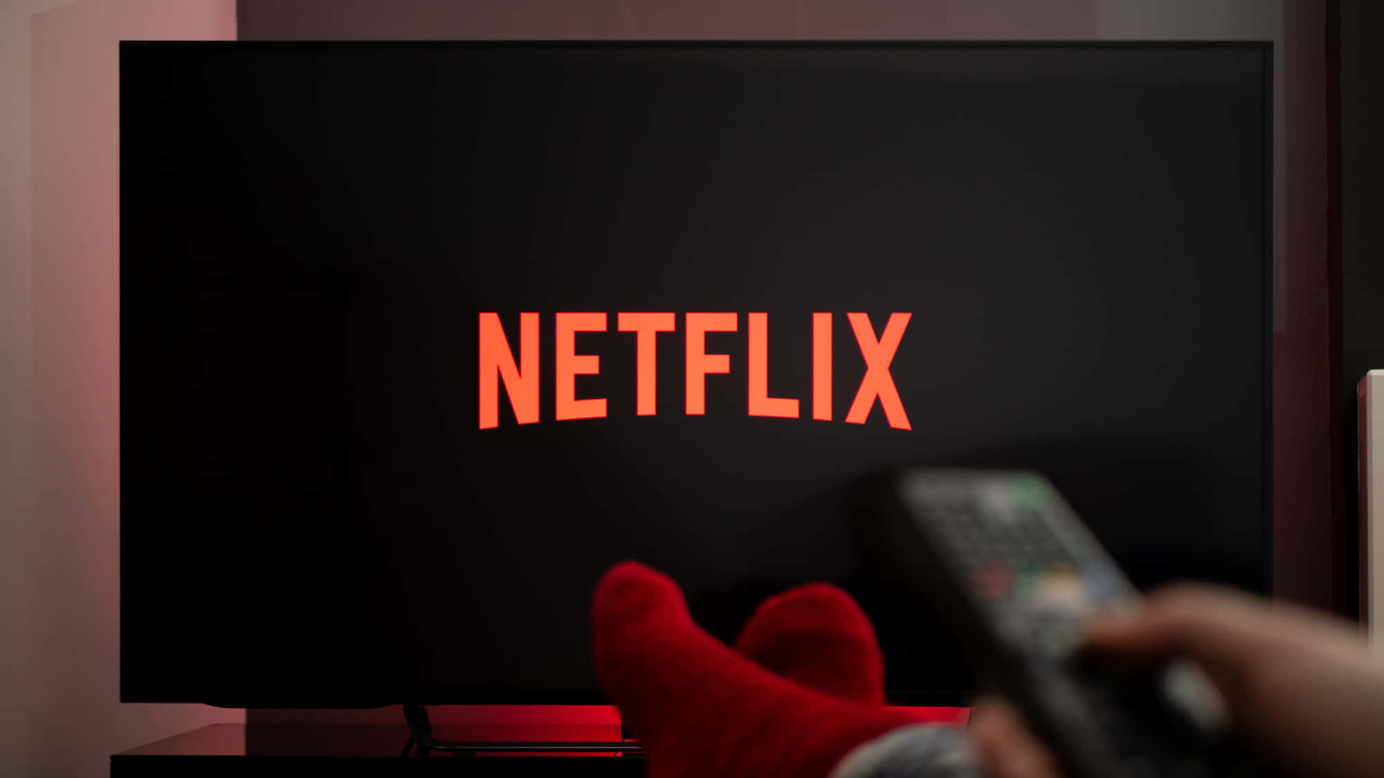 Netflix 桌布 壁紙 — 一隻手持遙控器的手指向一台電視，電視在昏暗的房間中顯示發光的橙色 Netflix 標誌。