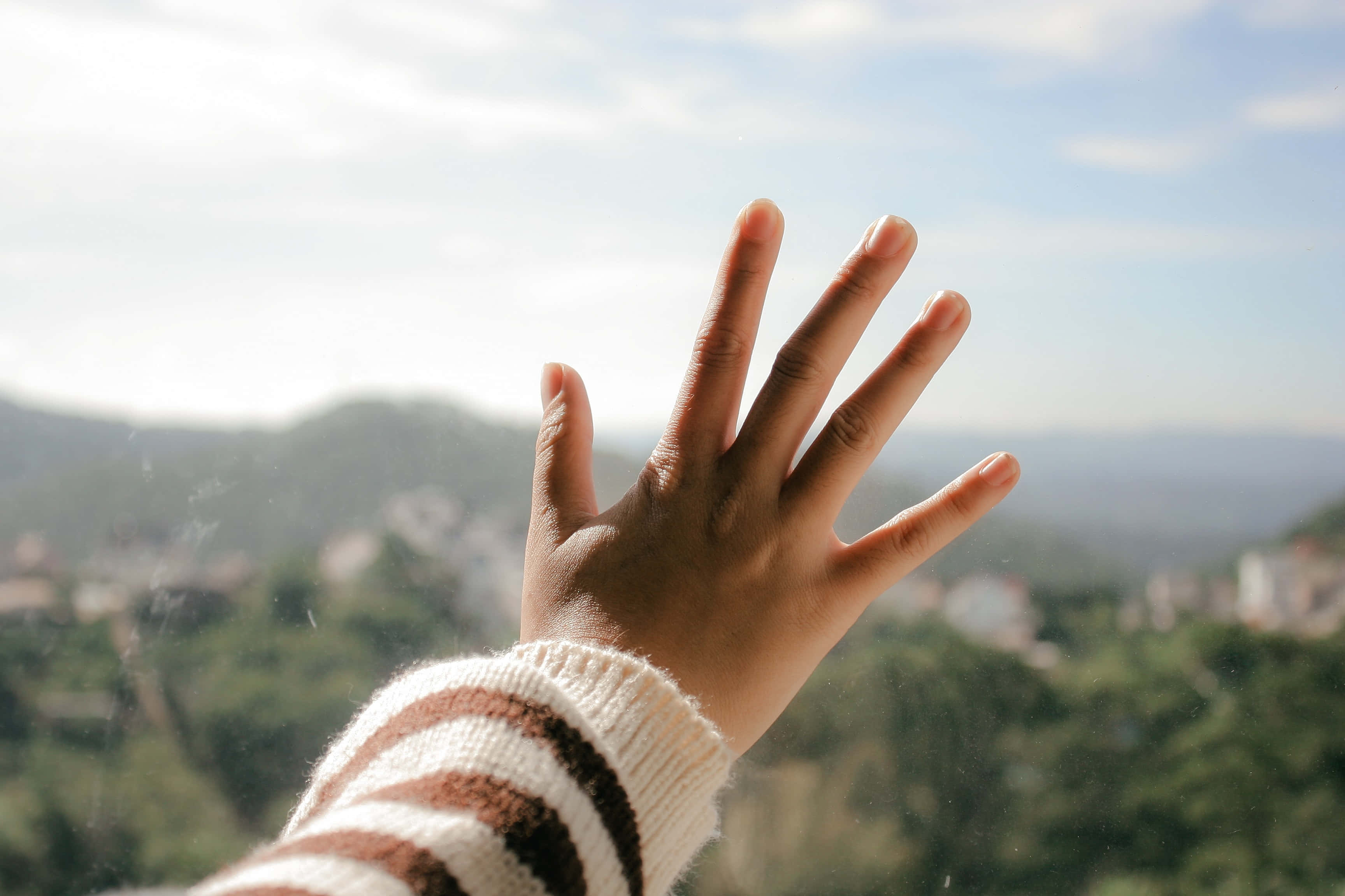 hands 桌布 壁紙 — 一隻手按在陽光照射的窗戶上，俯瞰著一個朦朧的綠色山谷，遠處有山丘和散落的建築物。