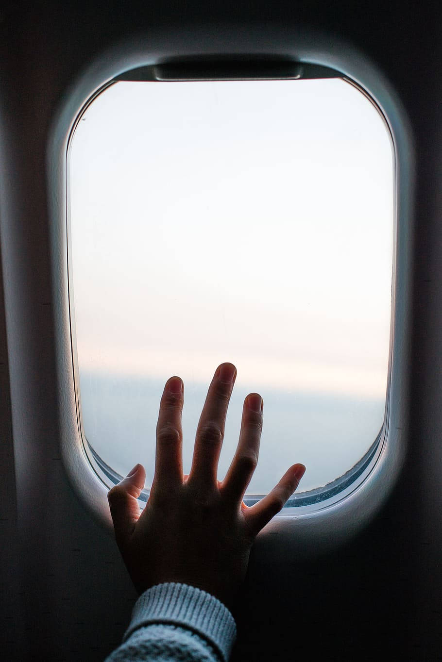 旅行 桌布 壁紙 — 一隻手按在飛機窗上，窗框映出黎明或黃昏時柔和的粉彩色天空。