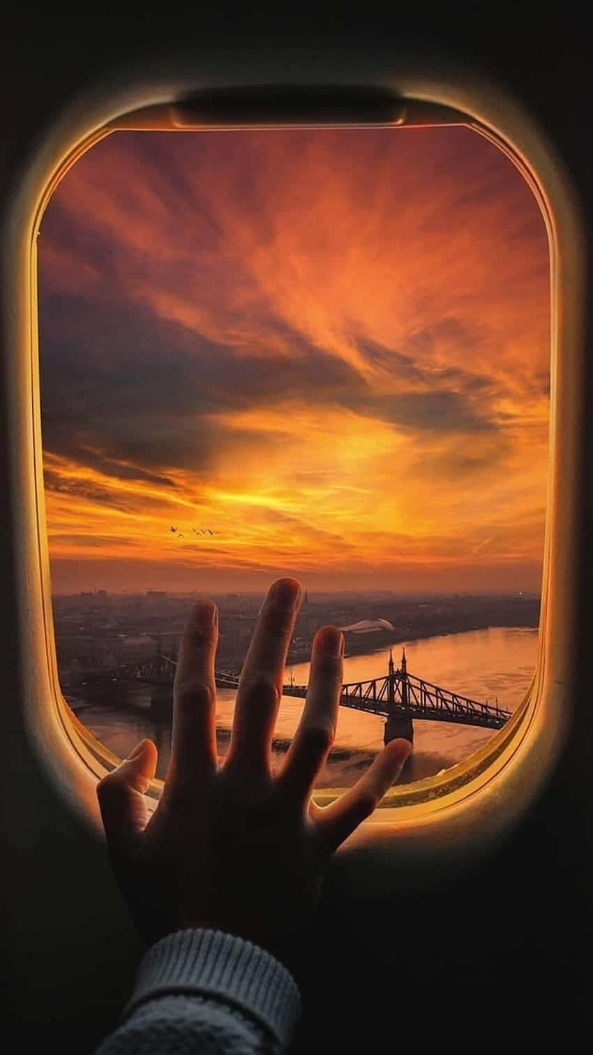 Airplane Window 桌布 壁紙 — 一隻手按壓在飛機窗戶上，框住河流和橋樑上方火熱的橙色和紅色日落。
