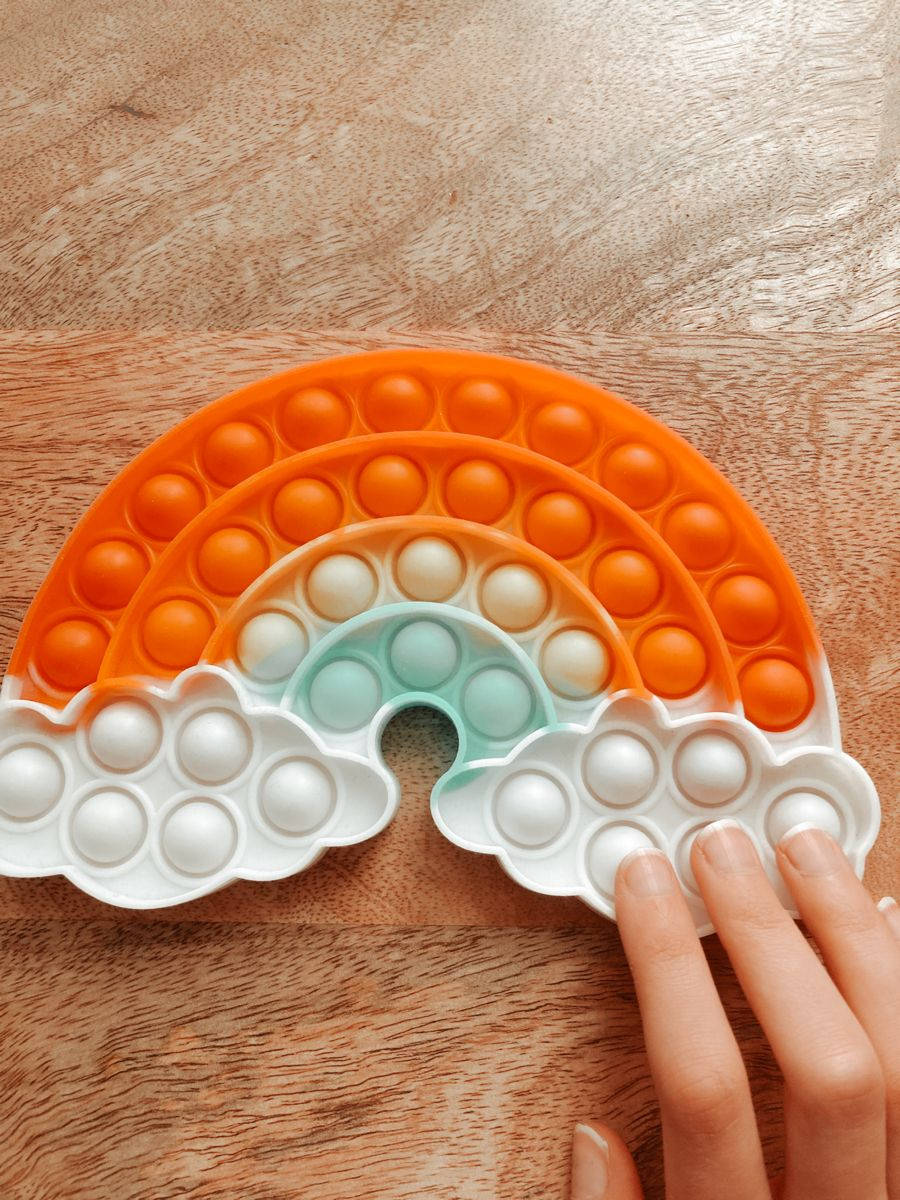 Fidget Toys 桌布 壁紙 — 一隻手按壓著放在溫暖木質表面上的彩虹形狀按壓玩具上的泡泡。
