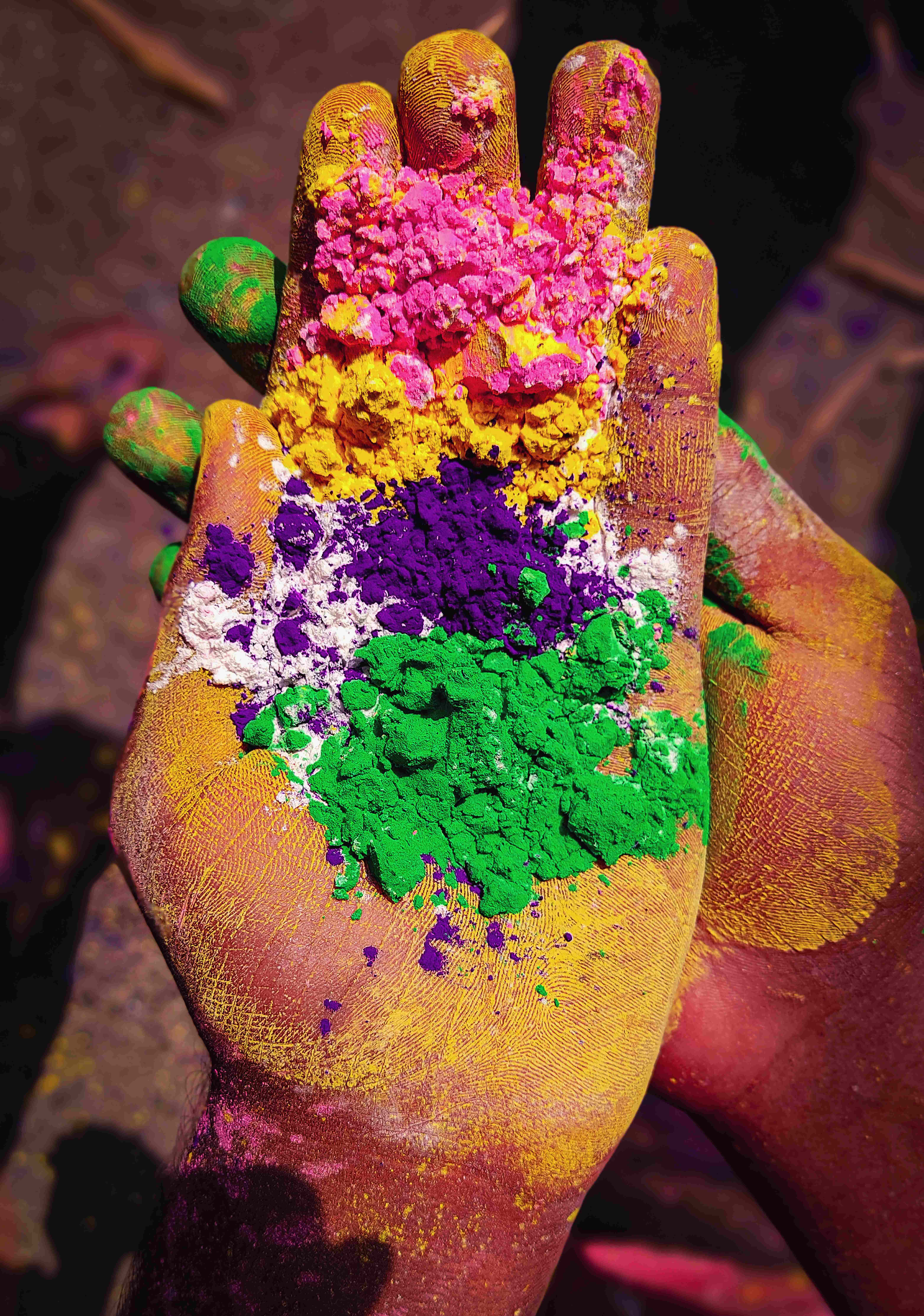 Holi 桌布 壁紙 — 一隻手捧著一堆色彩繽紛的粉末，包括粉紅、黃、紫、綠和白色，背景為深色。