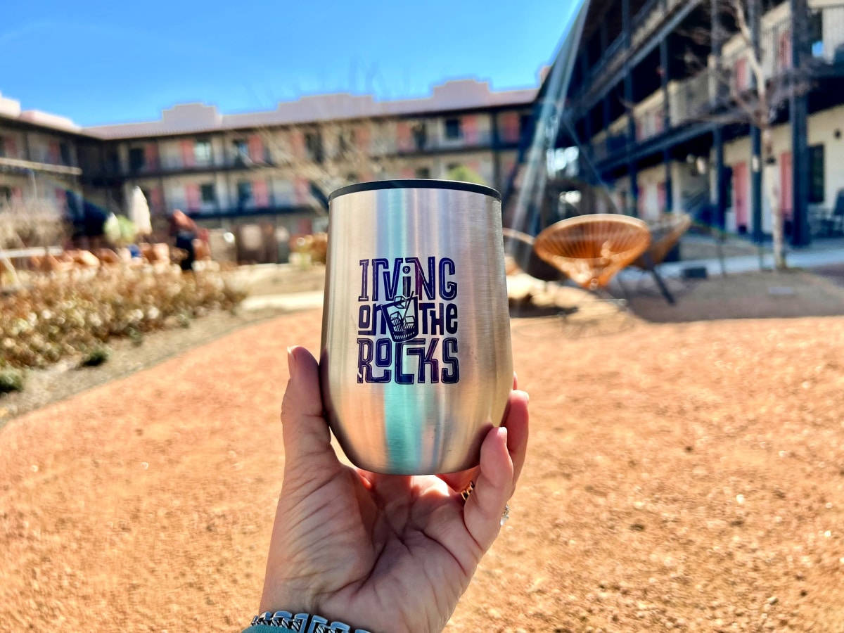 美國 桌布 壁紙 — 一隻手握住一個不鏽鋼杯，上面有「Irving on the Rocks」字樣，背景是陽光明媚的庭院，有建築物和乾燥的景觀。