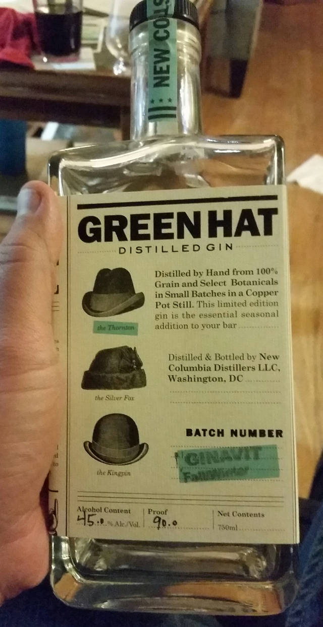 Gin 桌布 壁紙 — 一隻手握住一個透明的Green Hat Distilled Gin酒瓶，上面有詳細標籤顯示三種帽款和蒸餾資訊。
