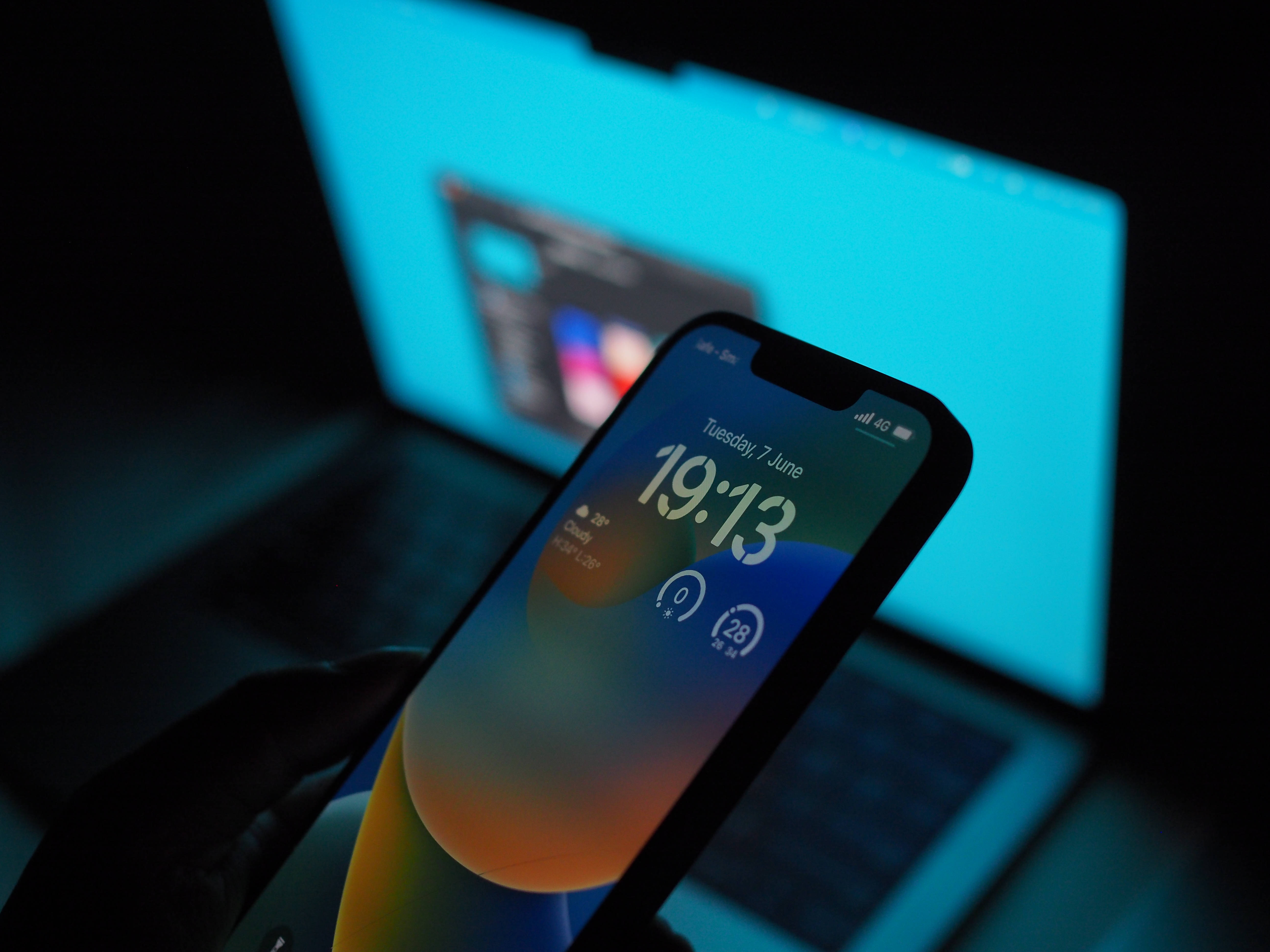 iOS 16 壁紙 桌布 壁紙 — 一隻手握住智慧型手機，顯示色彩繽紛的漸層桌布，帶有時間和天氣，背景是昏暗光線中發光的藍色筆記型電腦。