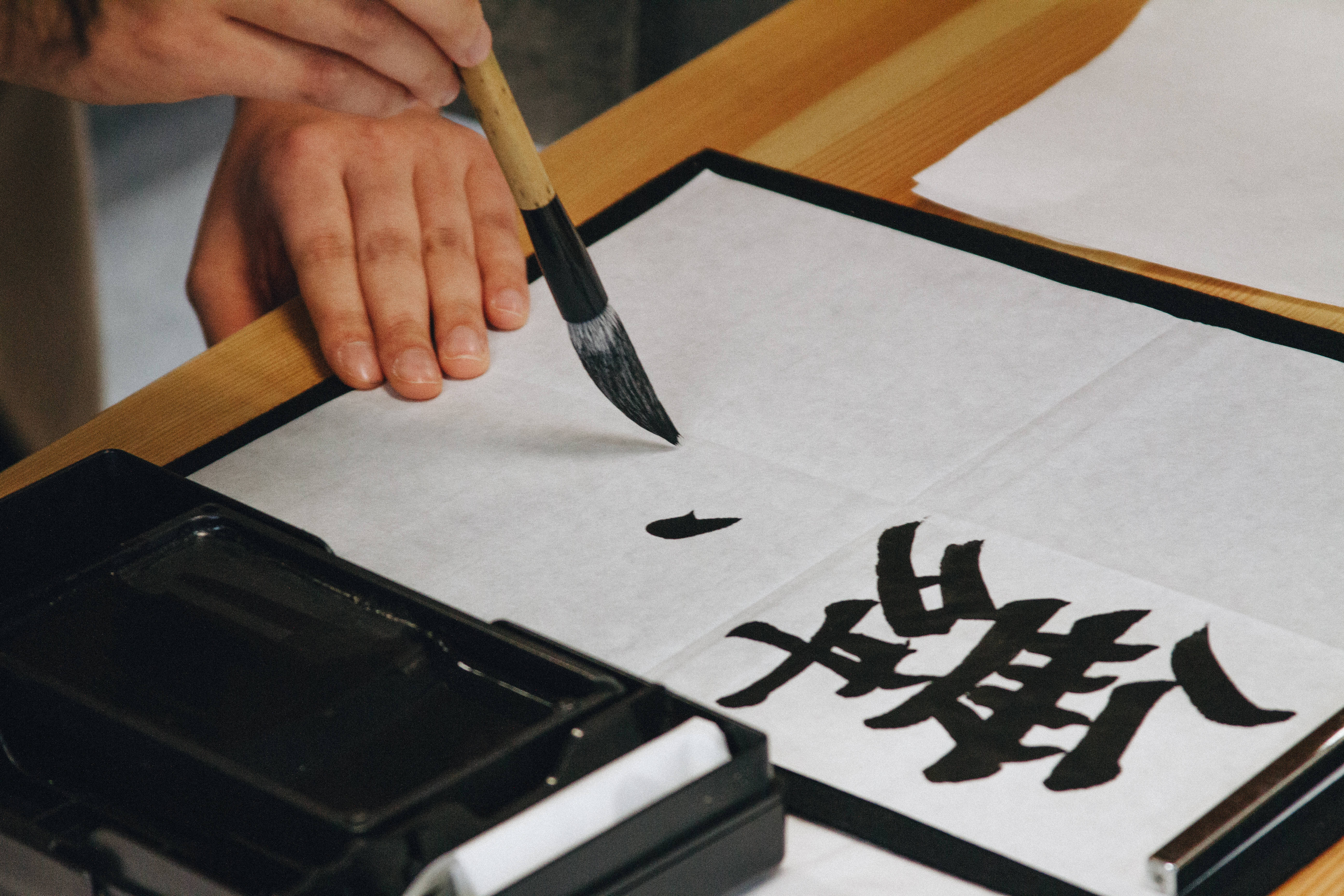 中文 桌布 壁紙 — 一隻手握住毛筆懸於宣紙之上，硯台旁粗獷的黑色書法筆觸在紙面上形成。