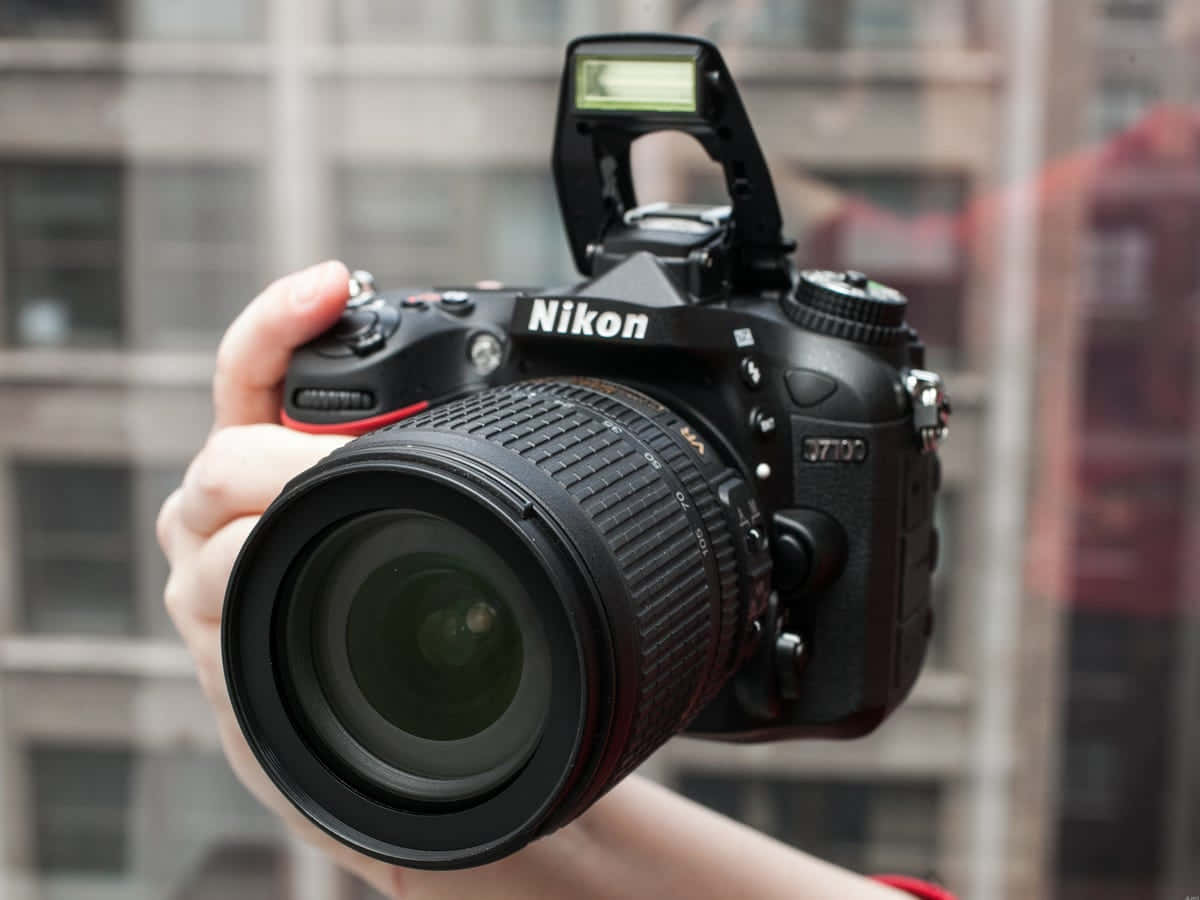 Dslr Camera 桌布 壁紙 — 一隻手握住黑色Nikon D7100 DSLR相機，鏡頭指向前方，背景是模糊的城市景觀。