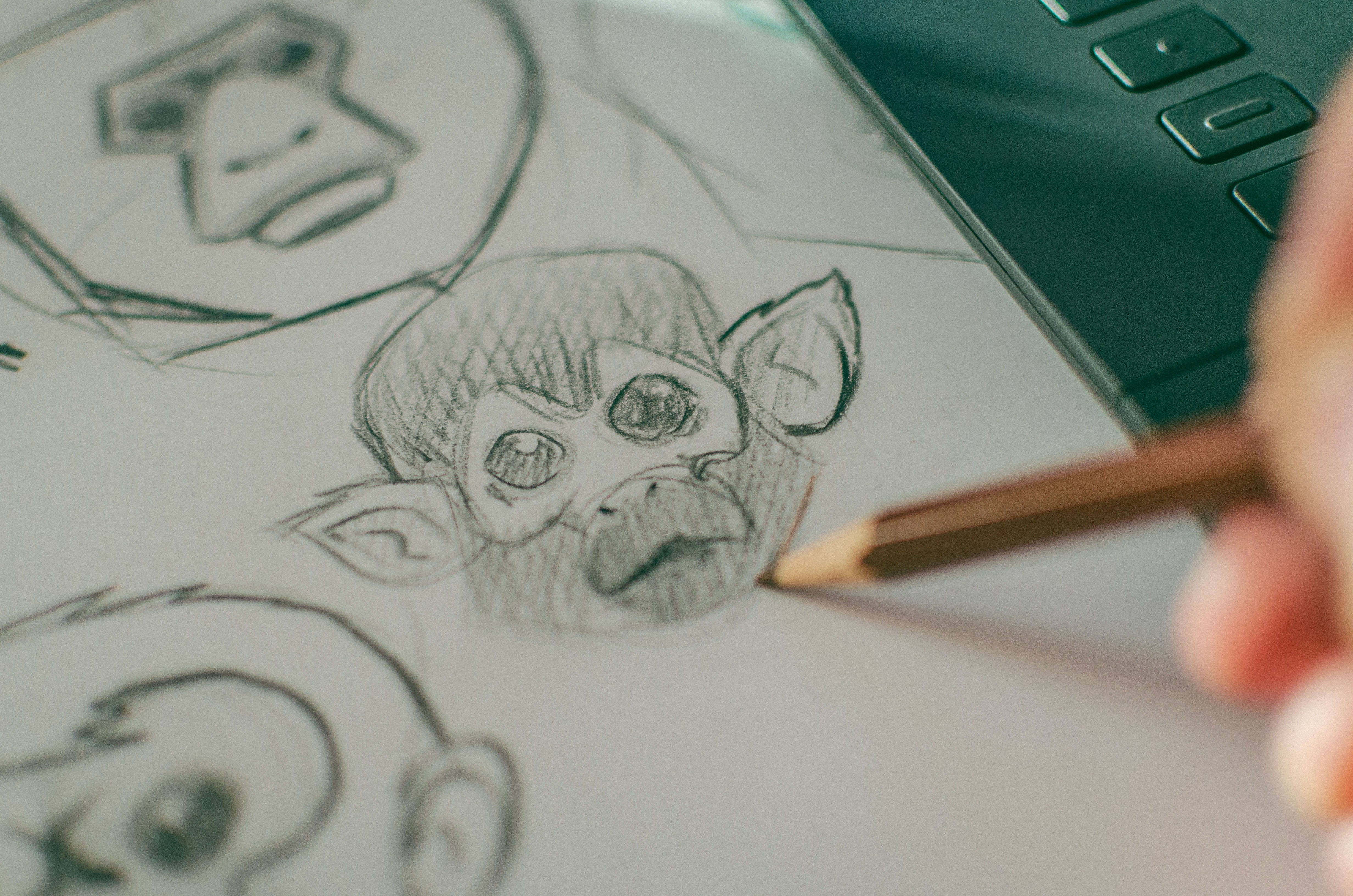 pencil 桌布 壁紙 — 一隻手正在圖形繪圖板旁邊的紙上，用鉛筆素描一個卡通猴子臉的詳細圖畫。