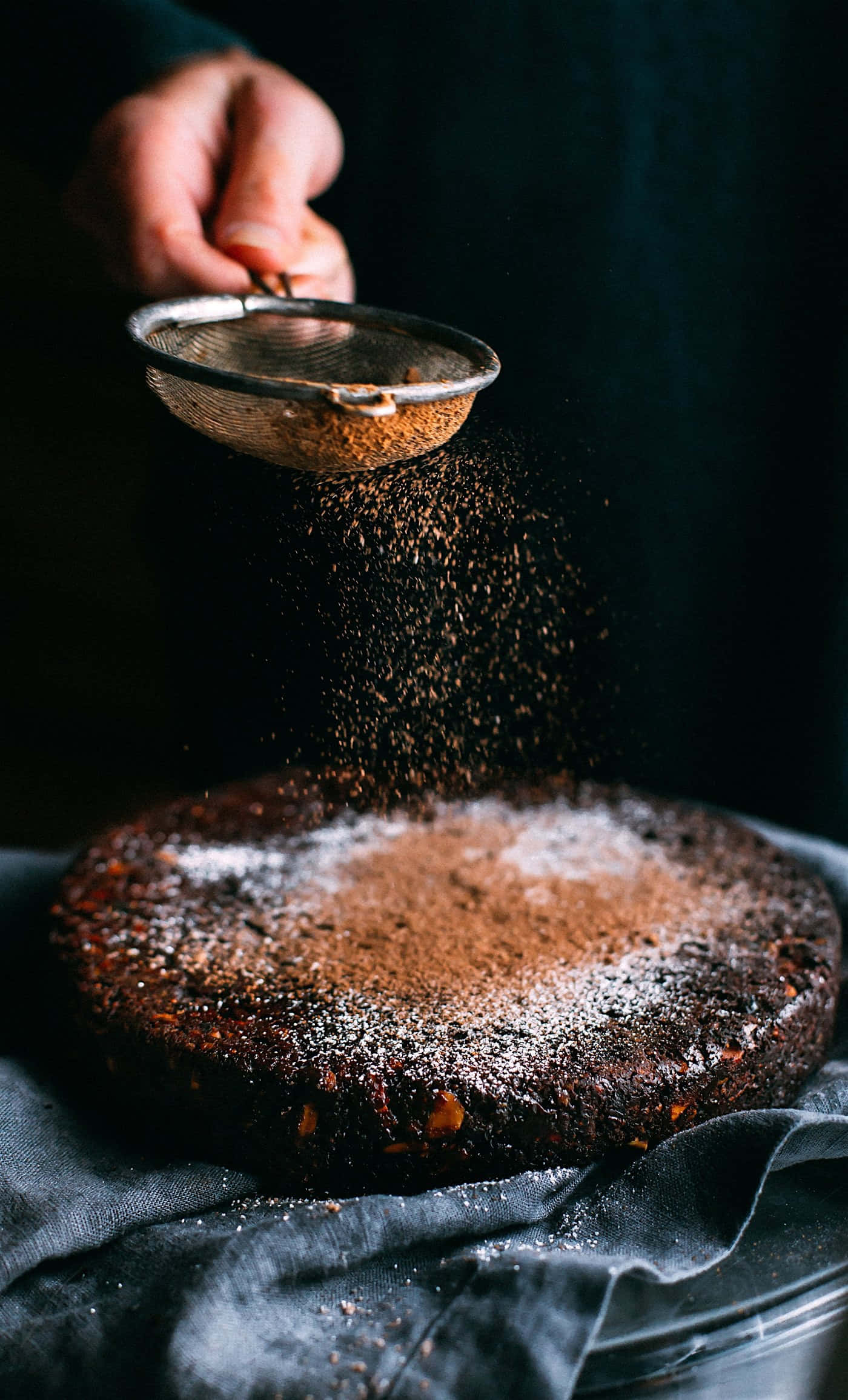 Baking 桌布 壁紙 — 一隻手正在將可可粉篩在一個放在深色布上的濃郁黑巧克力蛋糕上。
