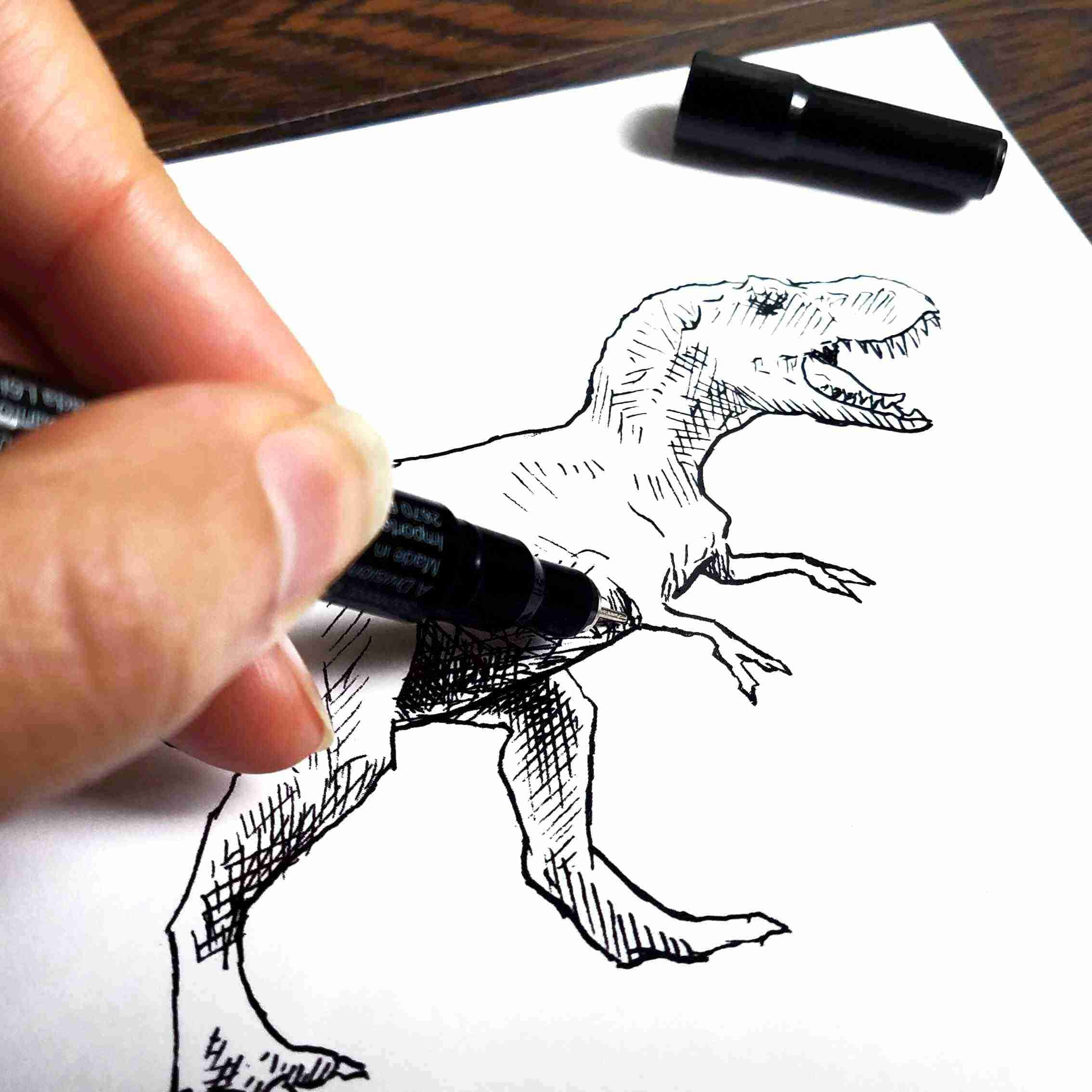 Dinosaur Drawing 桌布 壁紙 — 一隻手正在白紙上用黑色墨水繪畫詳細的暴龍素描，附近有一個標記筆蓋。