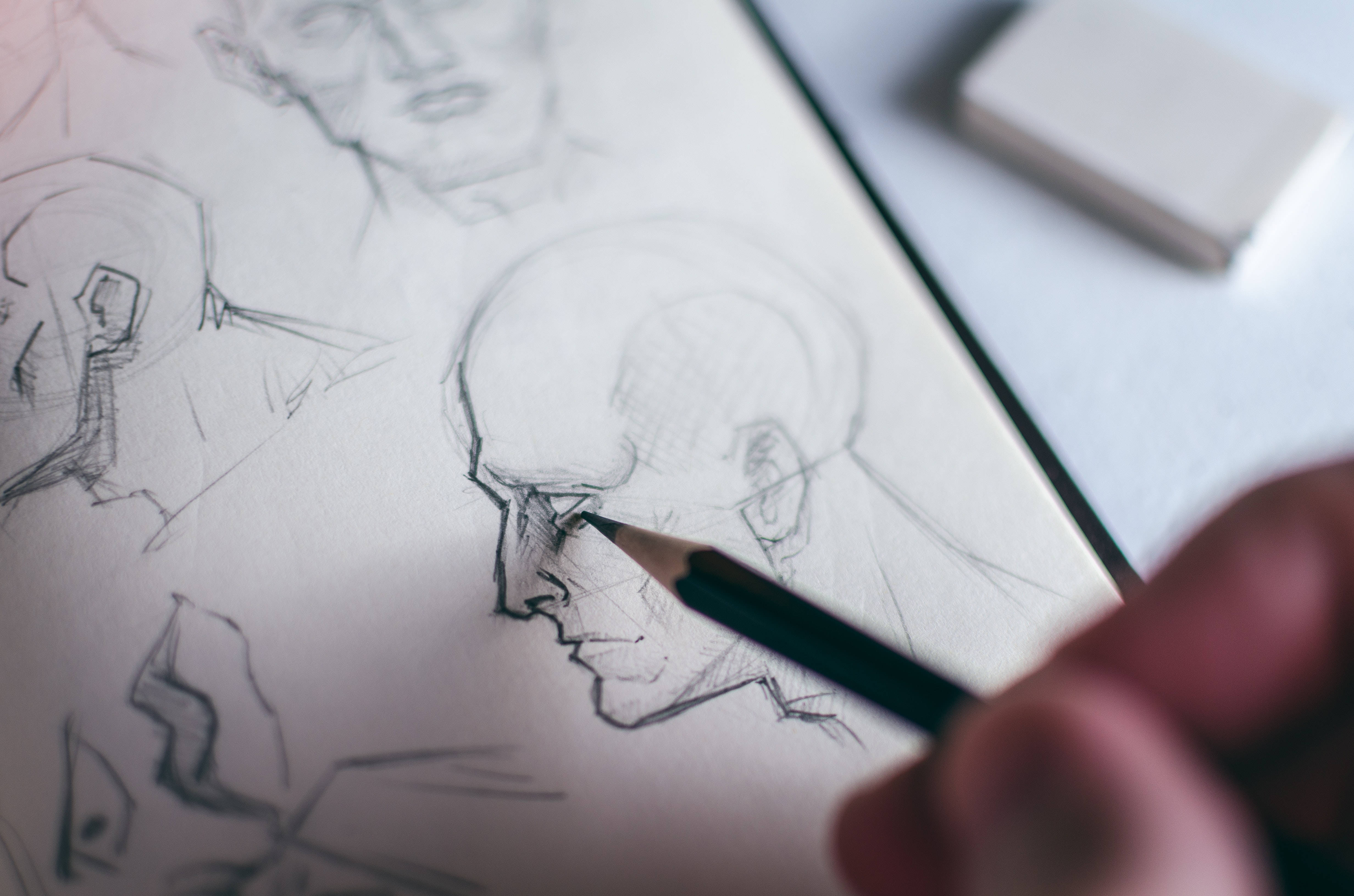 pencil 桌布 壁紙 — 一隻手正在紙上素描一個光頭男人臉部的詳細鉛筆肖像，周圍有其他粗略的草圖。