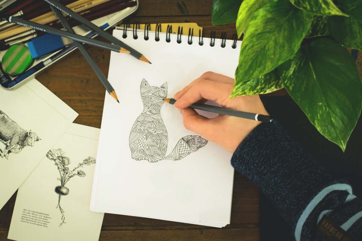Cat Drawing 桌布 壁紙 — 一隻手正在螺旋筆記本上繪畫一隻細節豐富的禪繞風格貓咪，周圍環繞著美術用品和一盆茂盛的綠色植物，放在木桌上。