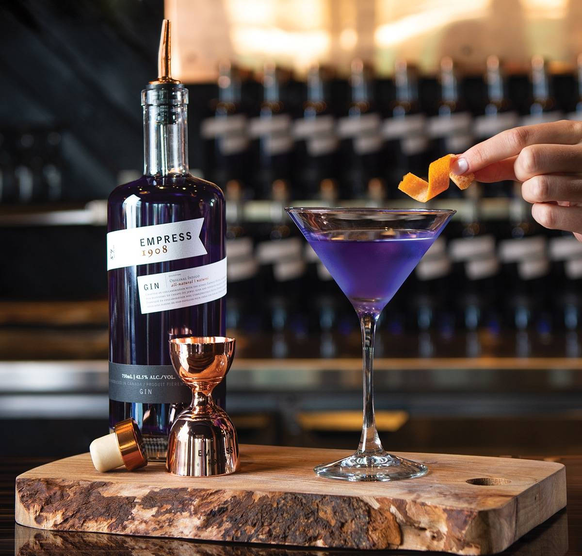martini 桌布 壁紙 — 一隻手正在裝飾一杯放在馬丁尼杯中的鮮豔紫色琴酒雞尾酒，旁邊有一瓶Empress 1908琴酒和一個銅製量酒器，放在質樸的木板上。