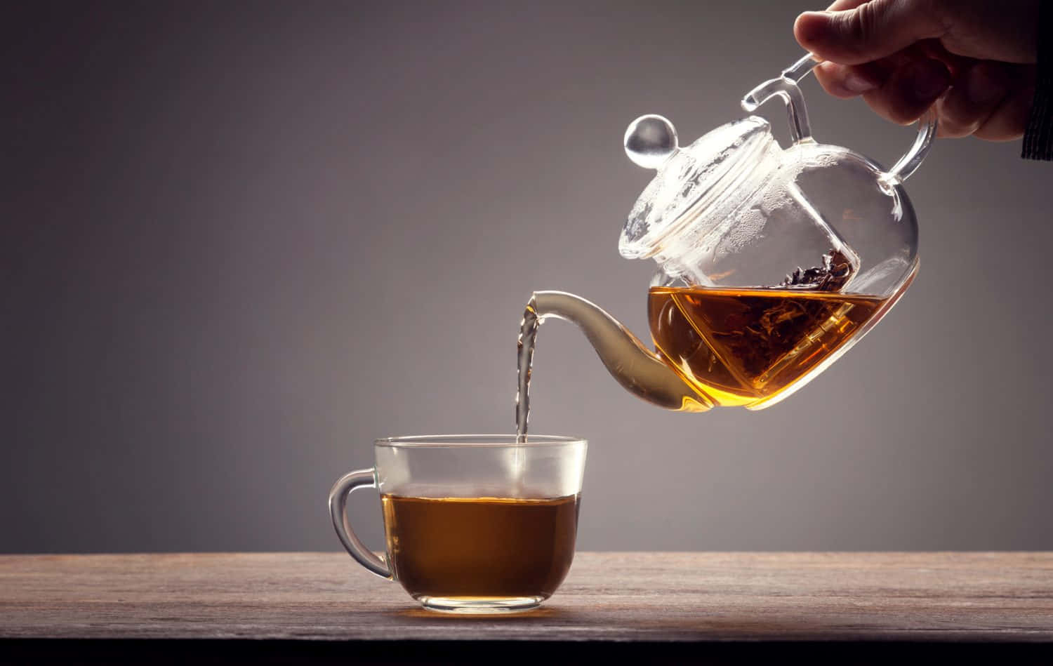 tea 桌布 壁紙 — 一隻手正將琥珀色茶從玻璃茶壺倒入木桌上的一個透明杯中，背景為柔和的灰色。