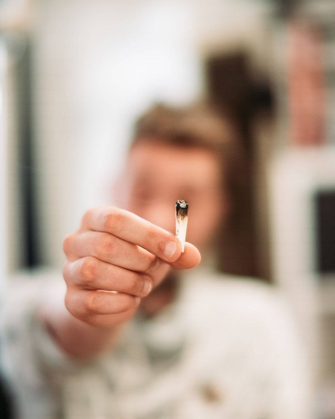 iPhone 攝影 桌布 壁紙 — 一隻手清晰對焦拿著一支點燃的大麻煙，背後有一個模糊的人站在柔和光線的室內空間。