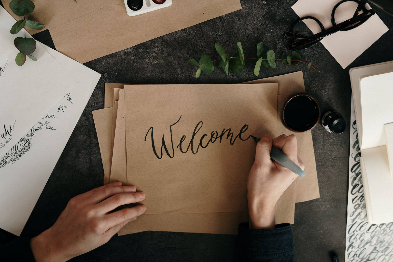 Welcome 桌布 壁紙 — 一隻手用優雅的黑色墨水在棕色紙上寫下「歡迎」一詞，置於深色紋理桌面上，周圍環繞著綠色植物和文具。