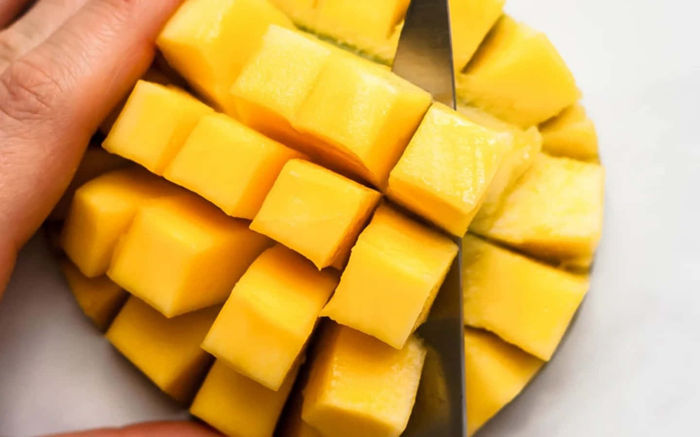 Mango 桌布 壁紙 — 一隻手用刀將成熟的芒果切成整齊的方塊，放在白色表面上。