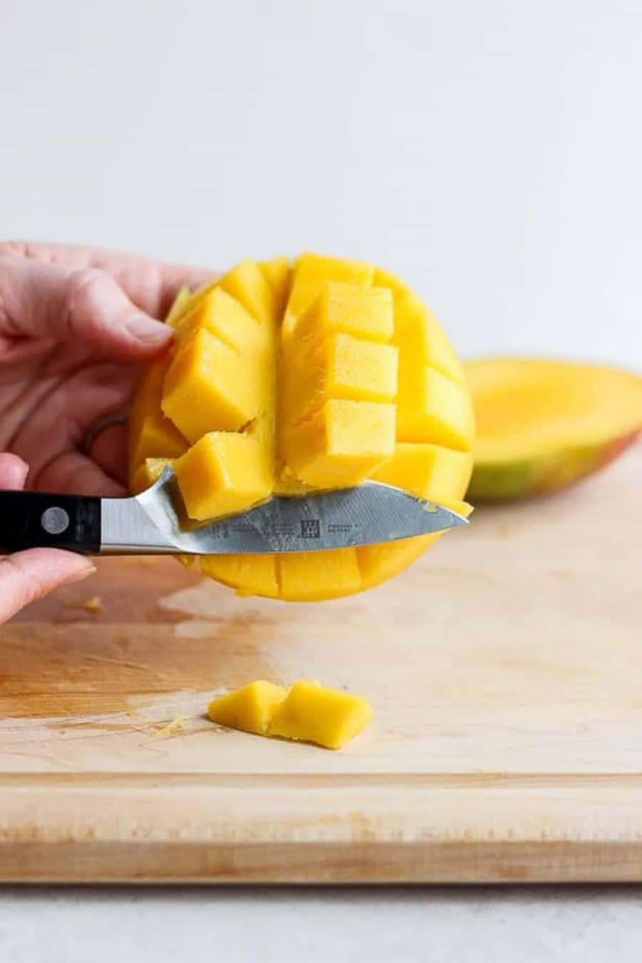 Mango 桌布 壁紙 — 一隻手用刀將成熟的黃芒果切成整齊的方塊，放在木製砧板上。