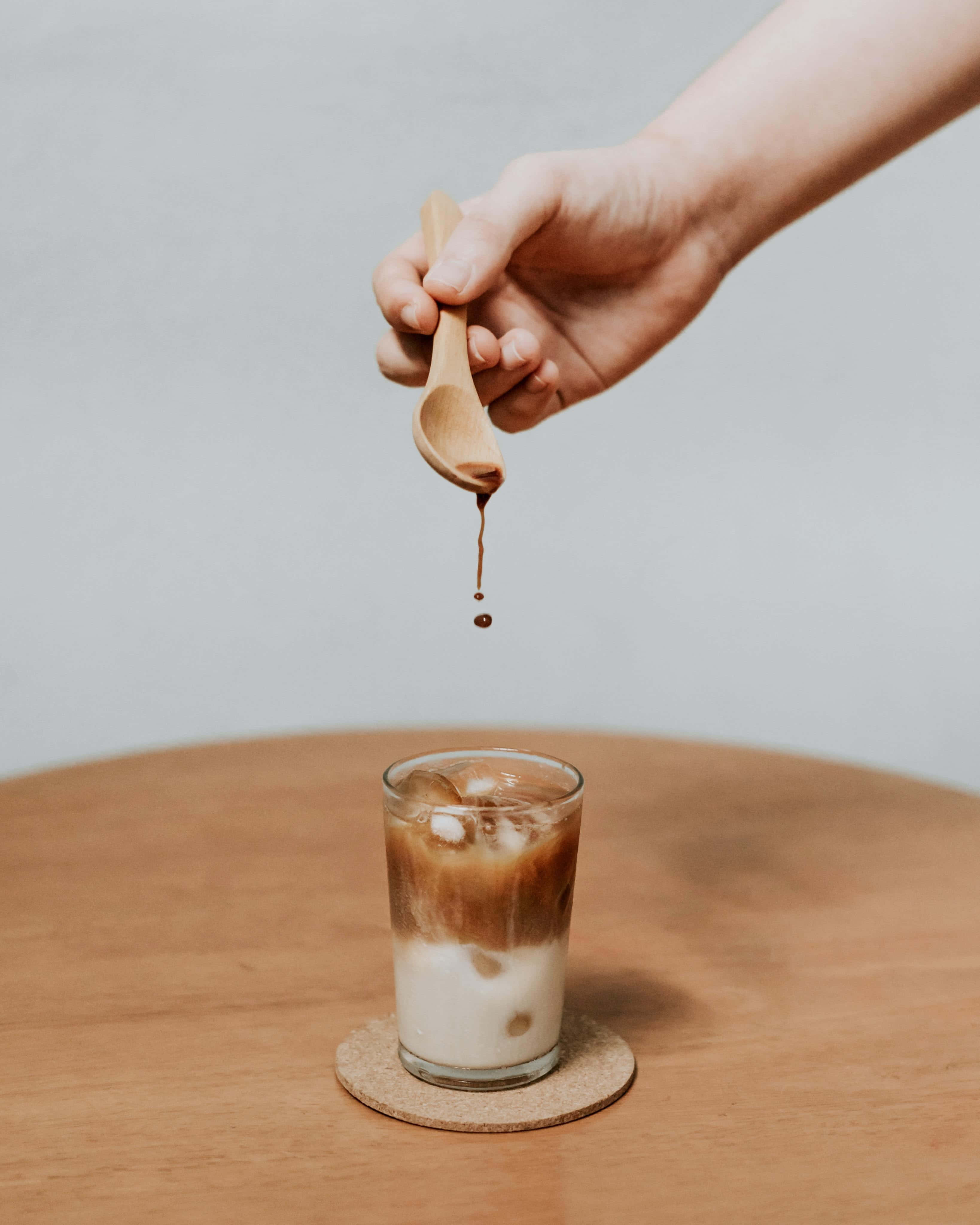 Iced Coffee 桌布 壁紙 — 一隻手用木匙將濃郁咖啡滴入木桌上的冰咖啡杯中。