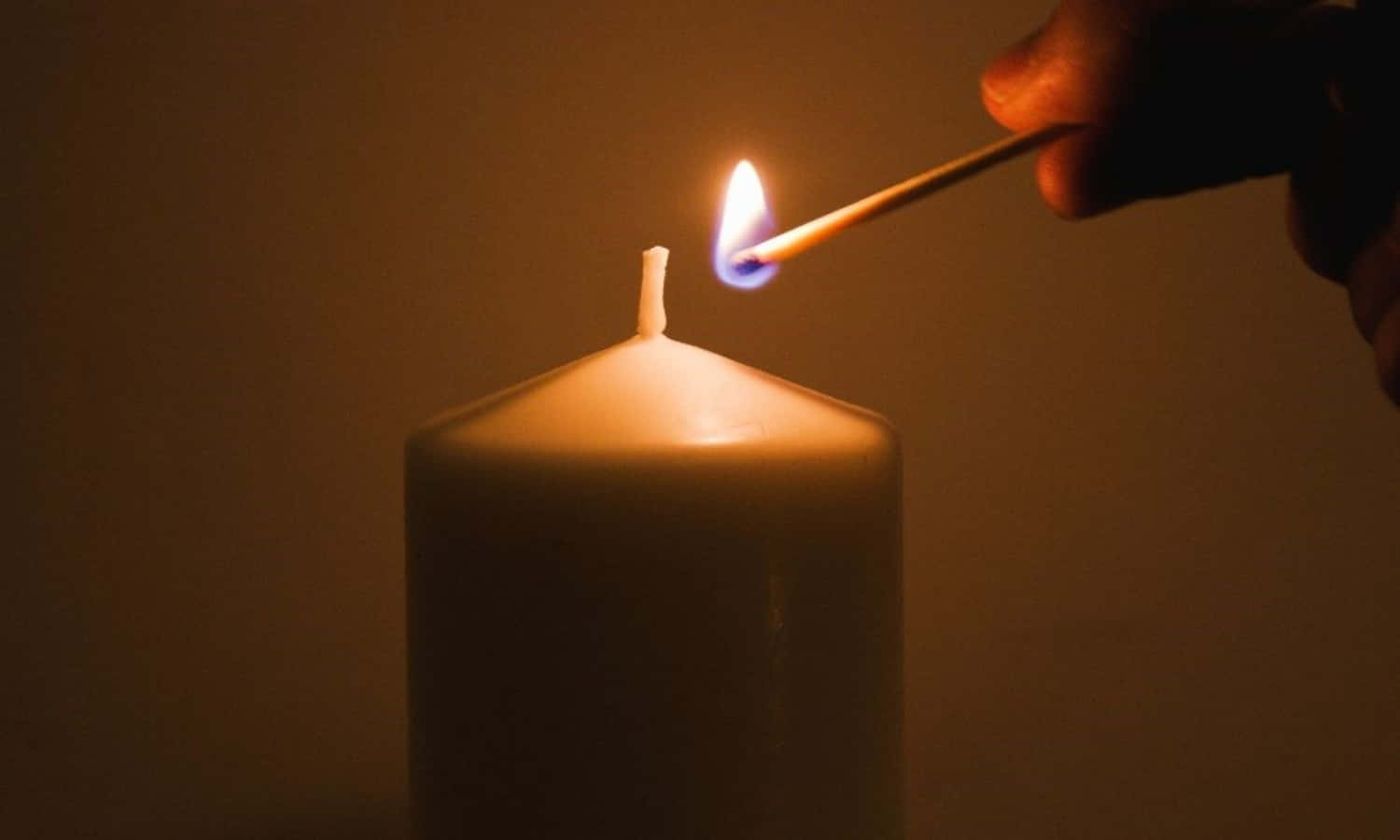 Candle Light 桌布 壁紙 — 一隻手用火柴點燃一支白色蠟燭，在昏暗溫暖的光線下投下柔和陰影。