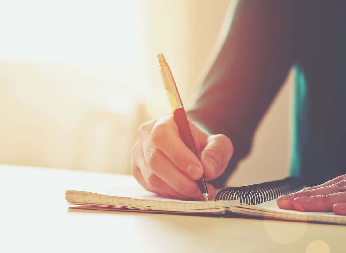 pencil 桌布 壁紙 — 一隻手用筆在筆記本上書寫，溫暖的陽光透過窗戶照射進來，營造出柔和的光暈。