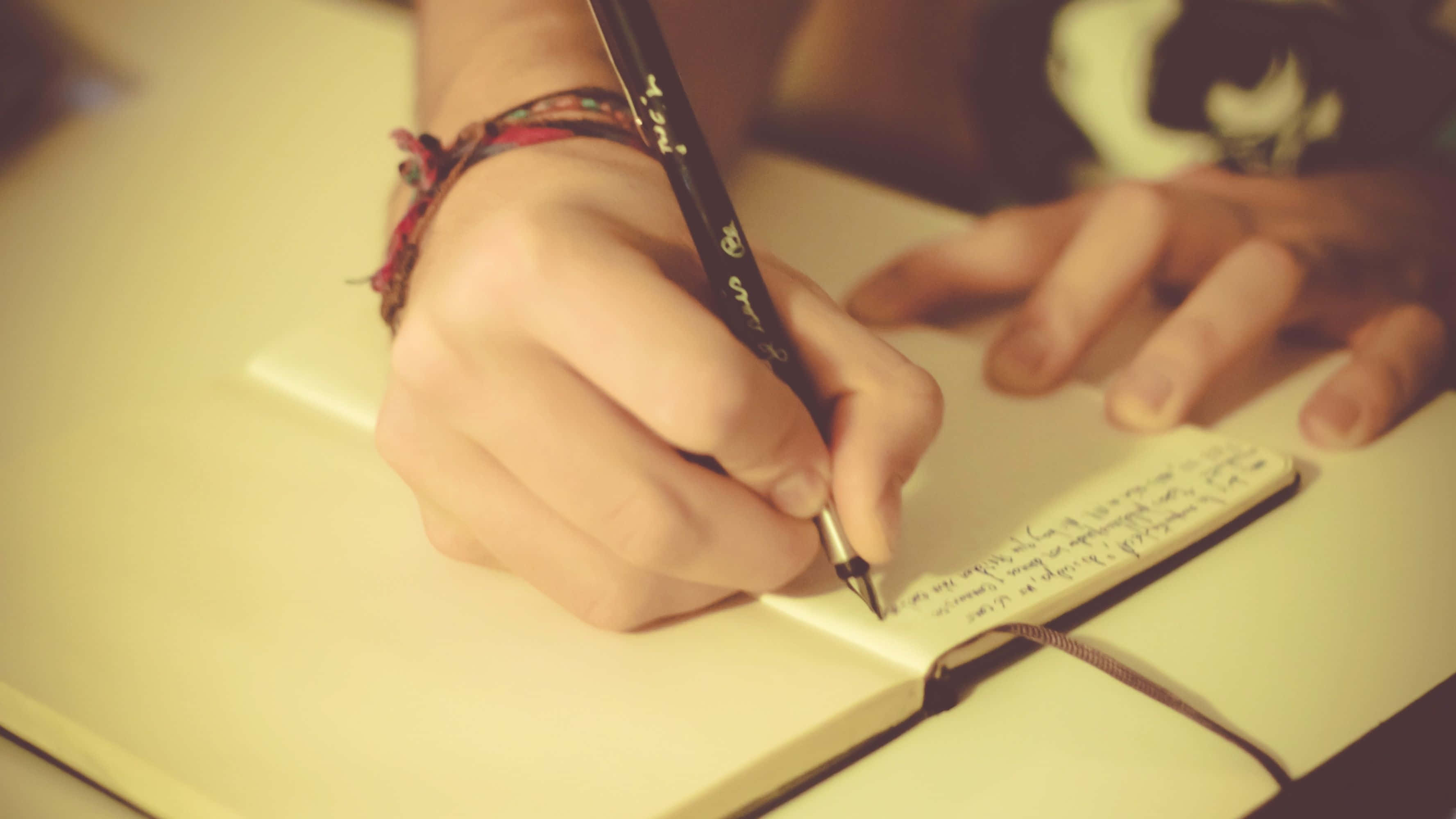 paper 桌布 壁紙 — 一隻手用筆在筆記本上書寫的特寫，戴著彩色手鐲，沐浴在溫暖的金色光線中。