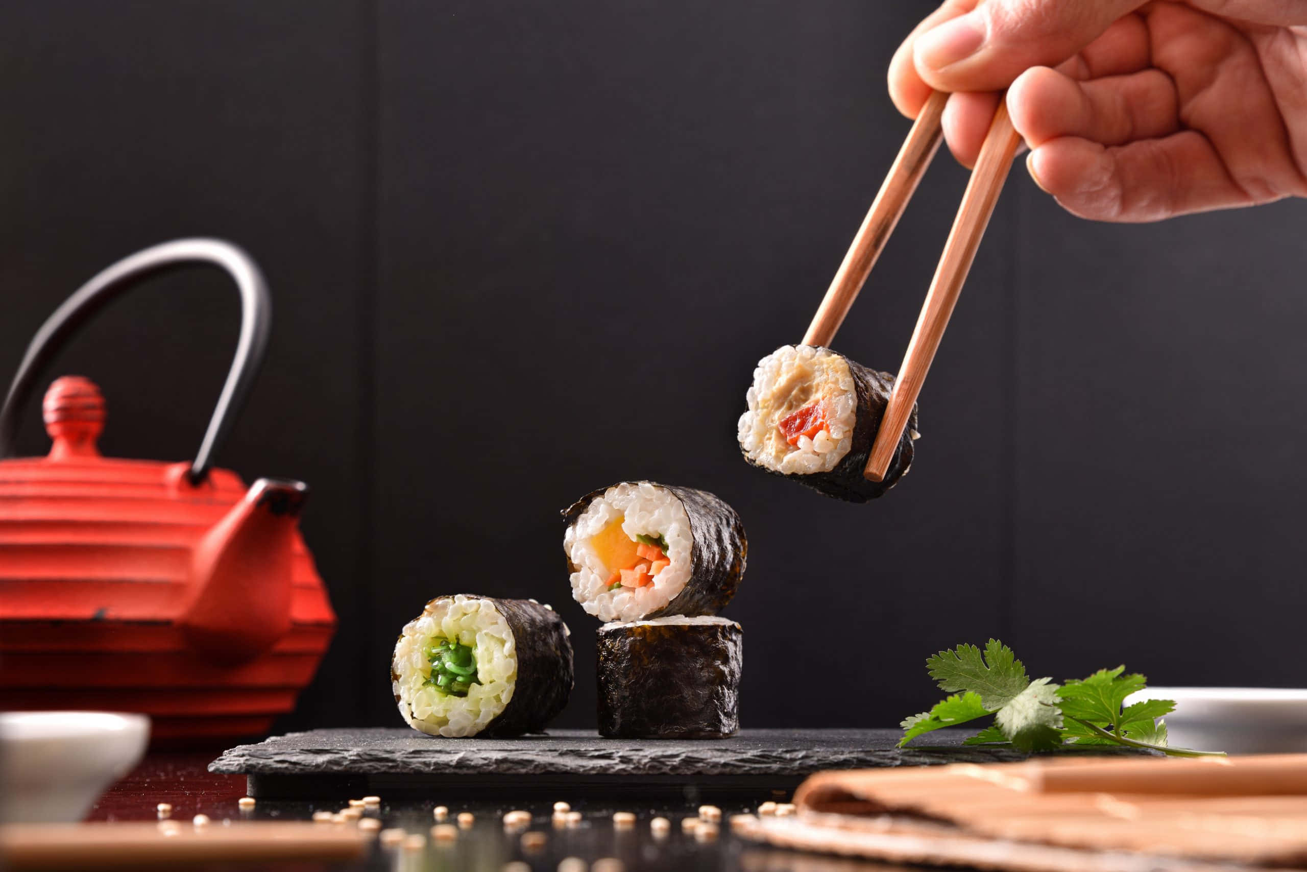 Sushi 桌布 壁紙 — 一隻手用筷子將壽司卷提起，放在一個石板盤上，背景有其他壽司卷和一個紅色茶壺。
