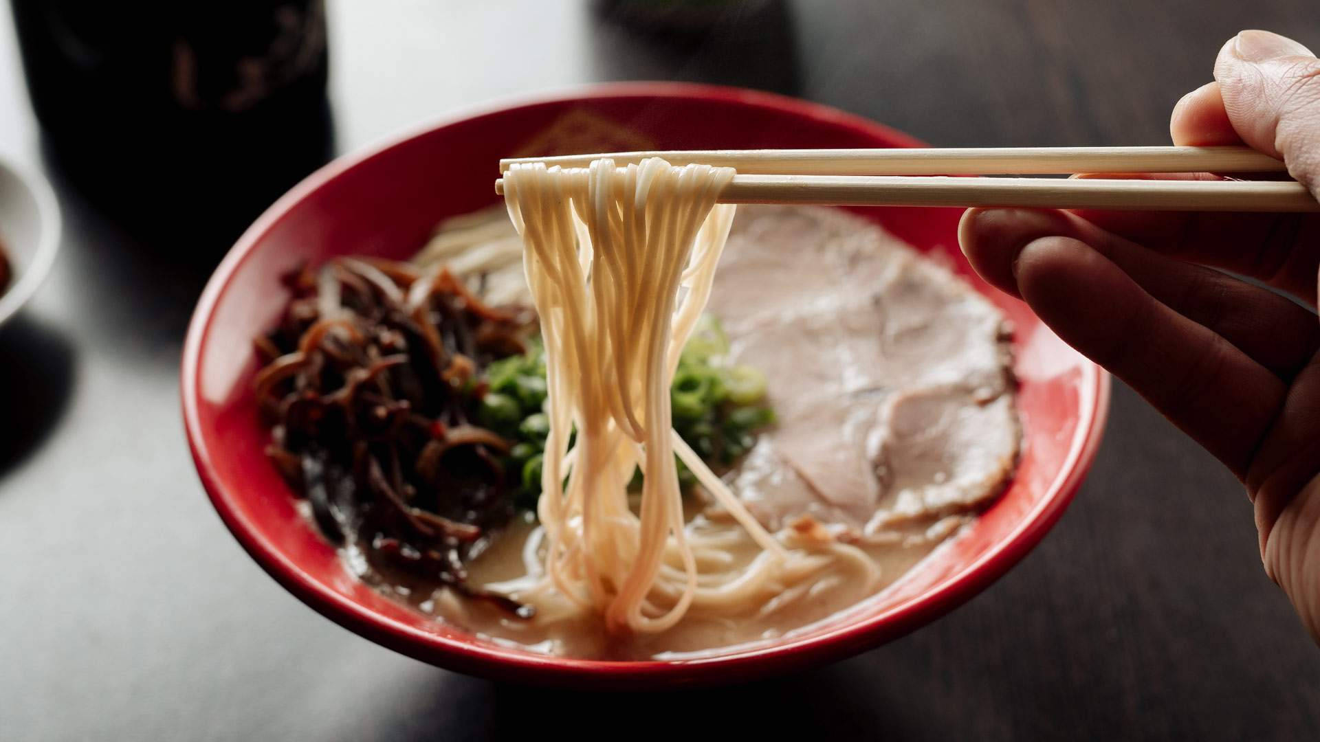 Ramen 桌布 壁紙 — 一隻手用筷子從一個充滿湯汁、豬肉和蔬菜的鮮紅色碗中夾起冒煙的拉麵。