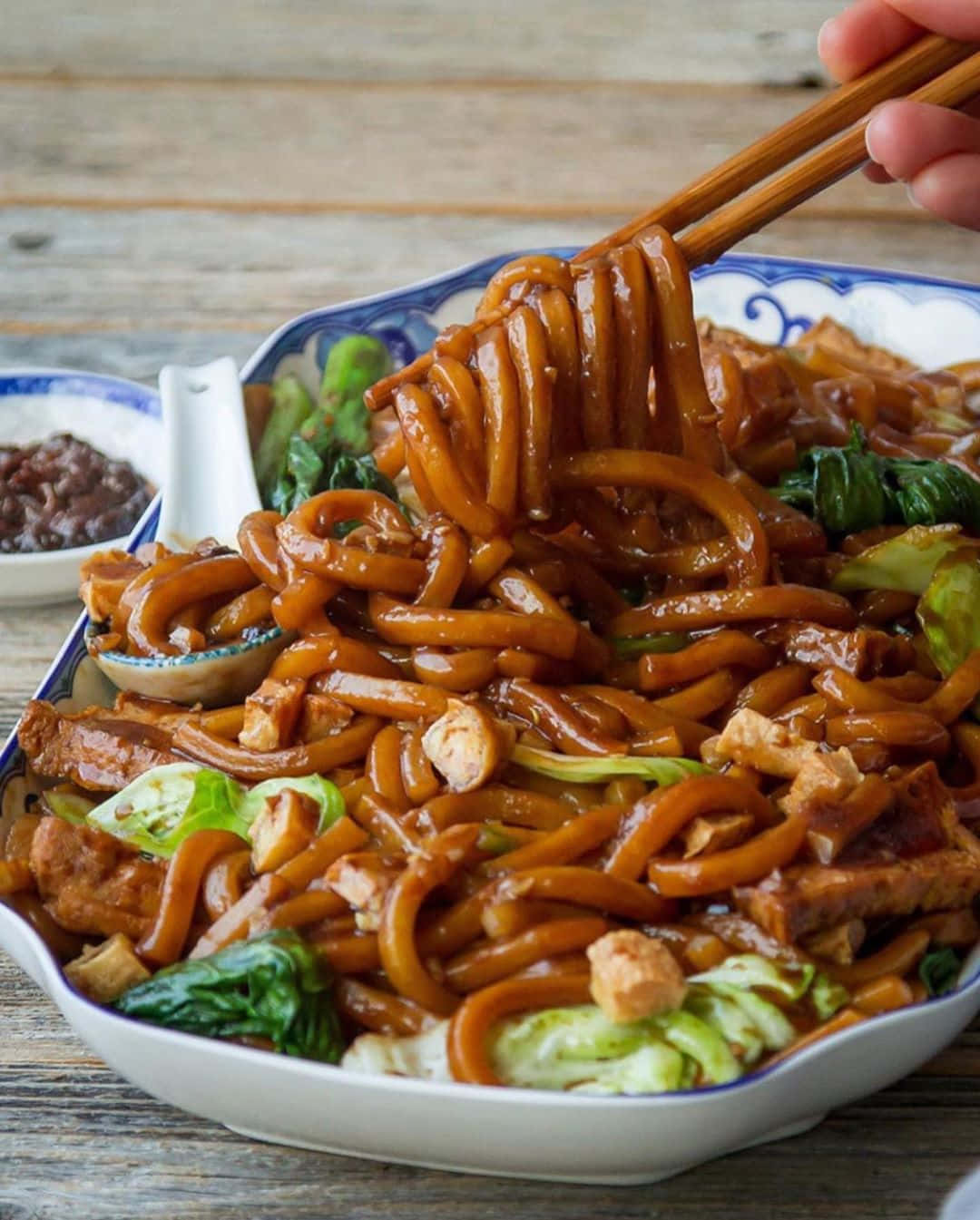 Food Photography 桌布 壁紙 — 一隻手用筷子從一個藍色花紋碗中夾起光滑的烏冬麵，碗裡有豆腐和小白菜，放在木桌上。