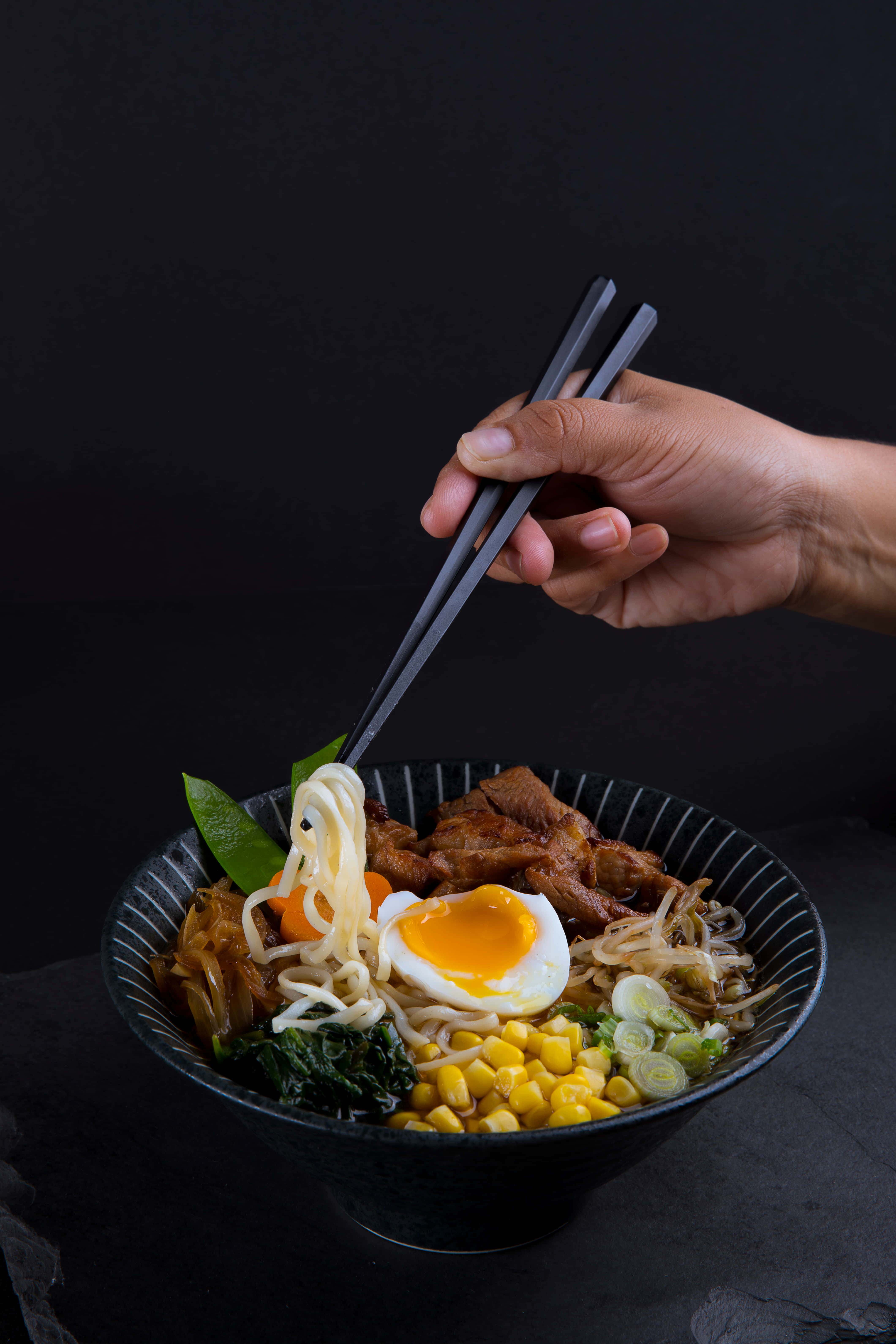 Noodles 桌布 壁紙 — 一隻手用筷子從一碗冒煙的拉麵中夾起麵條，上面有半熟蛋、玉米和青菜，背景深色。
