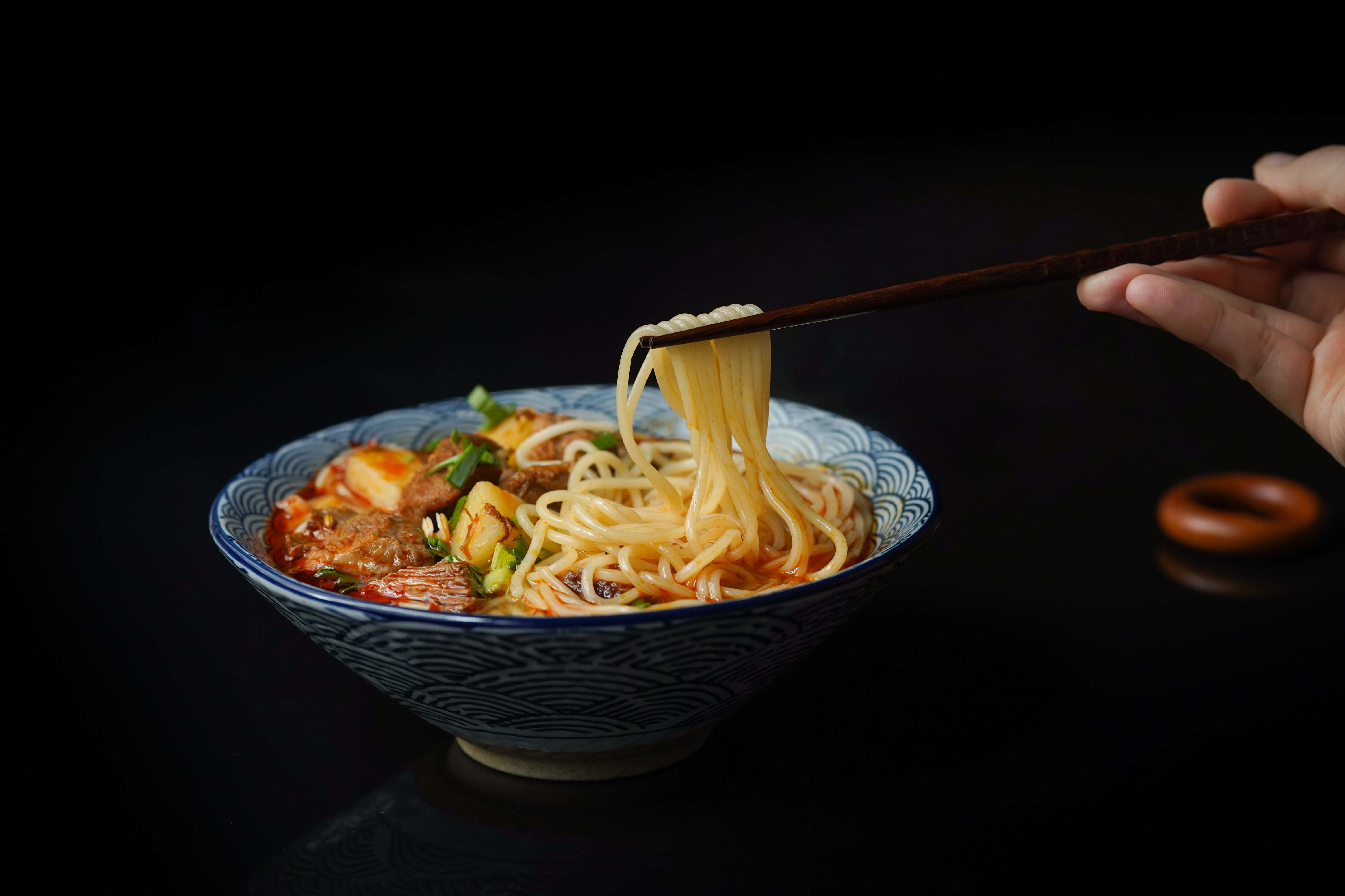 Food Photography 桌布 壁紙 — 一隻手用筷子從藍色花紋碗中夾起冒煙的麵條，背景為深色。