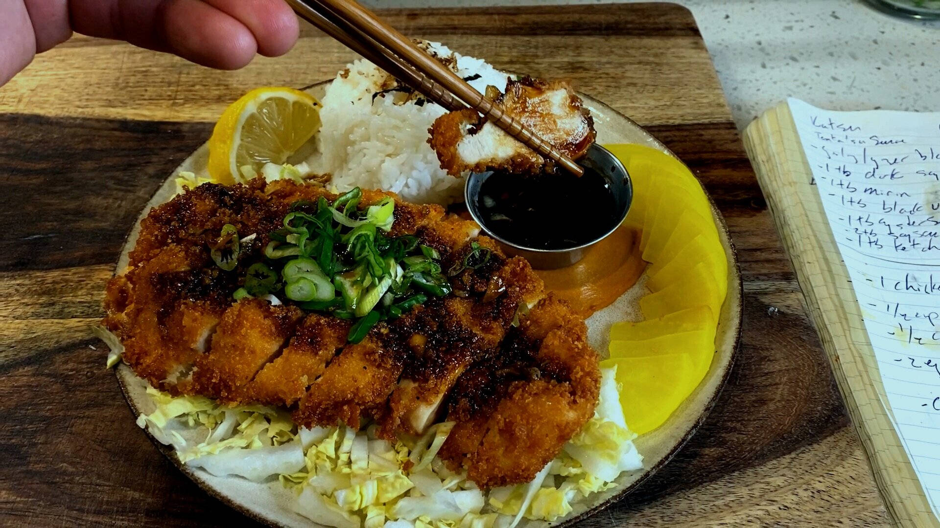 Food Photography 桌布 壁紙 — 一隻手用筷子從鋪有米飯、高麗菜和檸檬角的盤子上夾起酥脆的炸豬排。