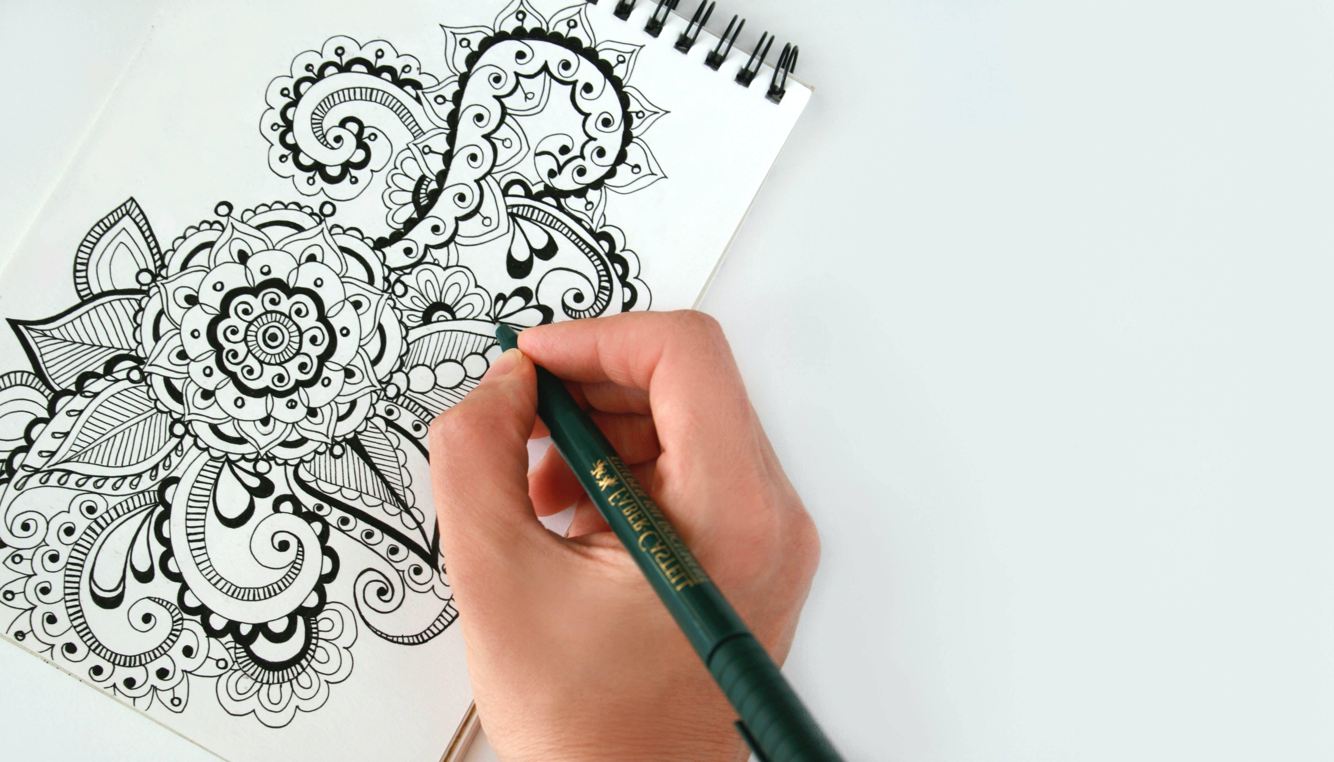 pencil 桌布 壁紙 — 一隻手用綠色筆在白色螺旋筆記本上繪製複雜的黑色曼陀羅圖案，背景為純白色。