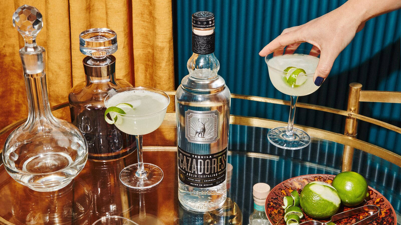 cocktail 桌布 壁紙 — 一隻手用萊姆裝飾瑪格麗特，而一瓶Cazadores龍舌蘭酒和雞尾酒杯在鏡面吧檯上閃閃發光。